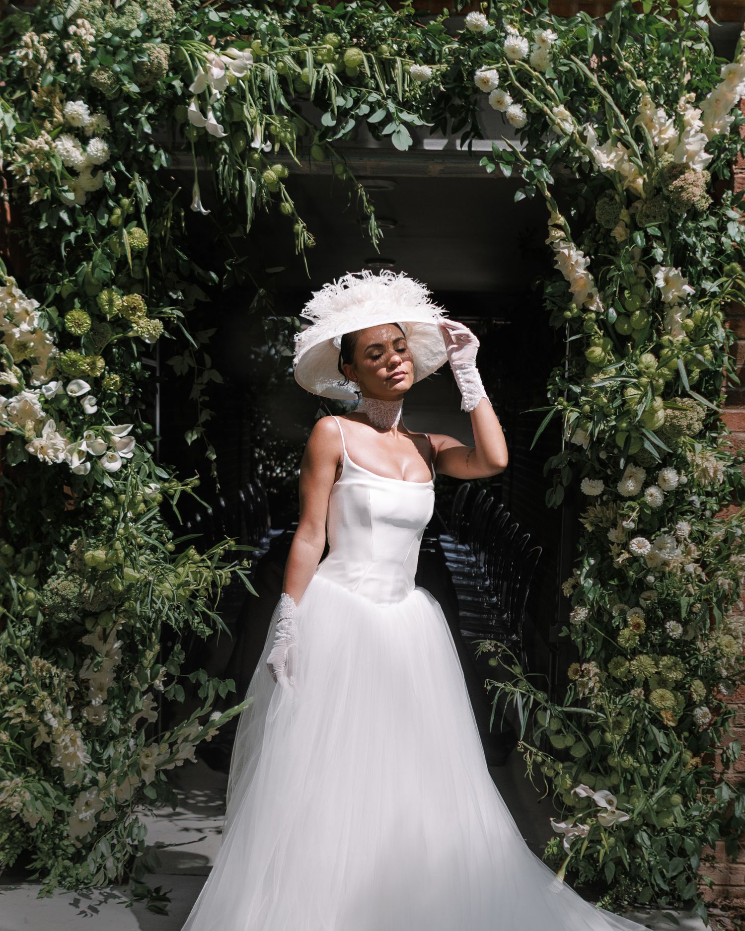 Bride in white tulle gown and wide-brim hat poses under floral arch with lush greenery.