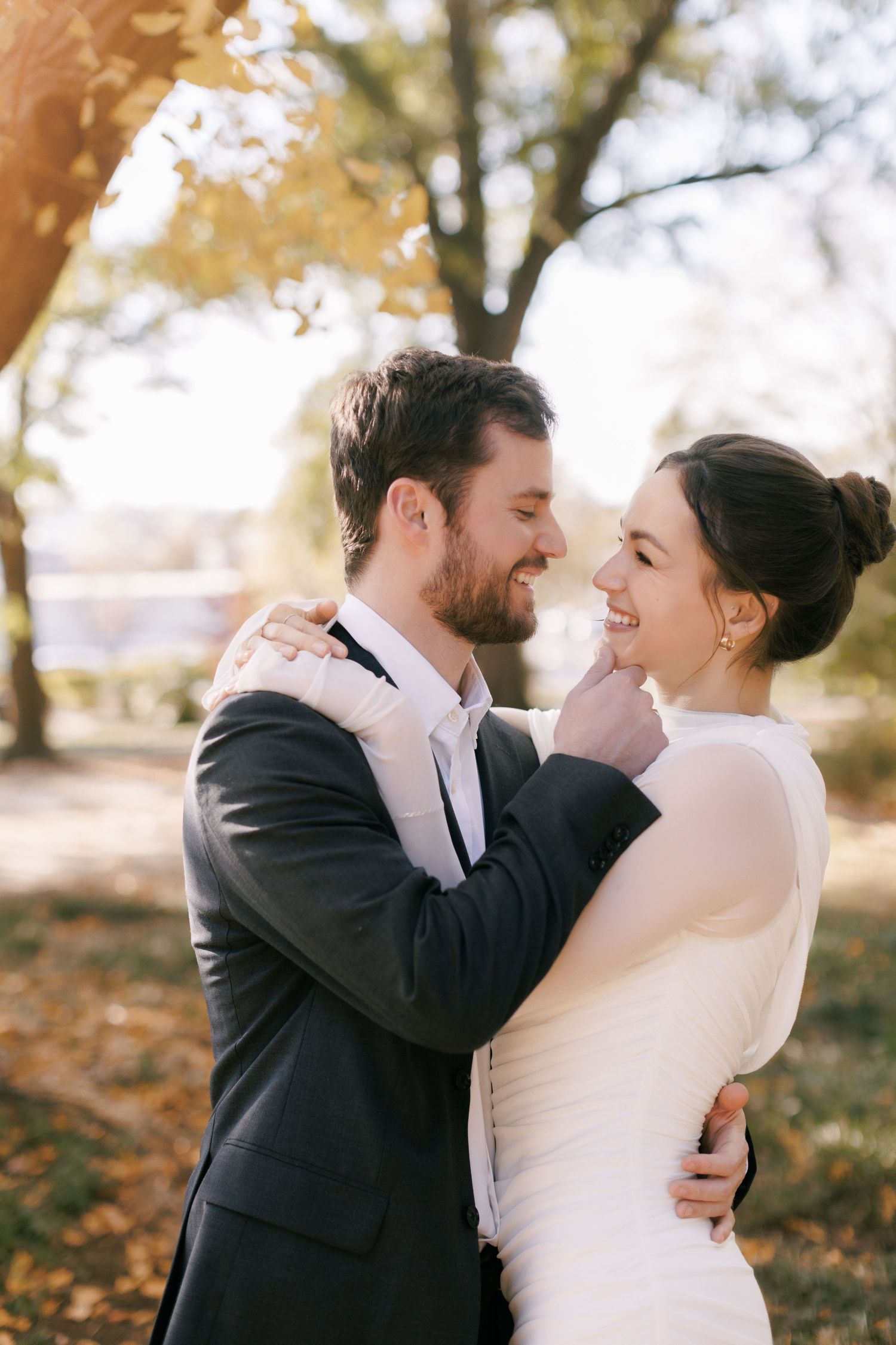 A smiling bride and groom embrace outdoors in an autumn park, sharing a romantic moment in wedding attire.