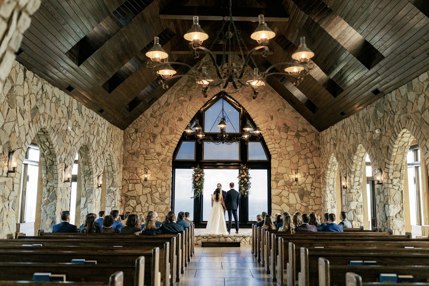 A rustic stone church interior with wooden beams and hanging lights creates a warm, intimate atmosphere for a wedding ceremony.
