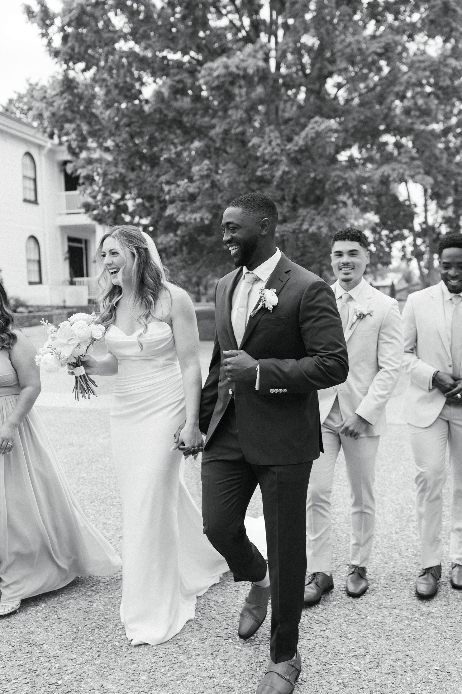 A joyful wedding party walks together outdoors with the newlyweds leading the group in black and white photography.