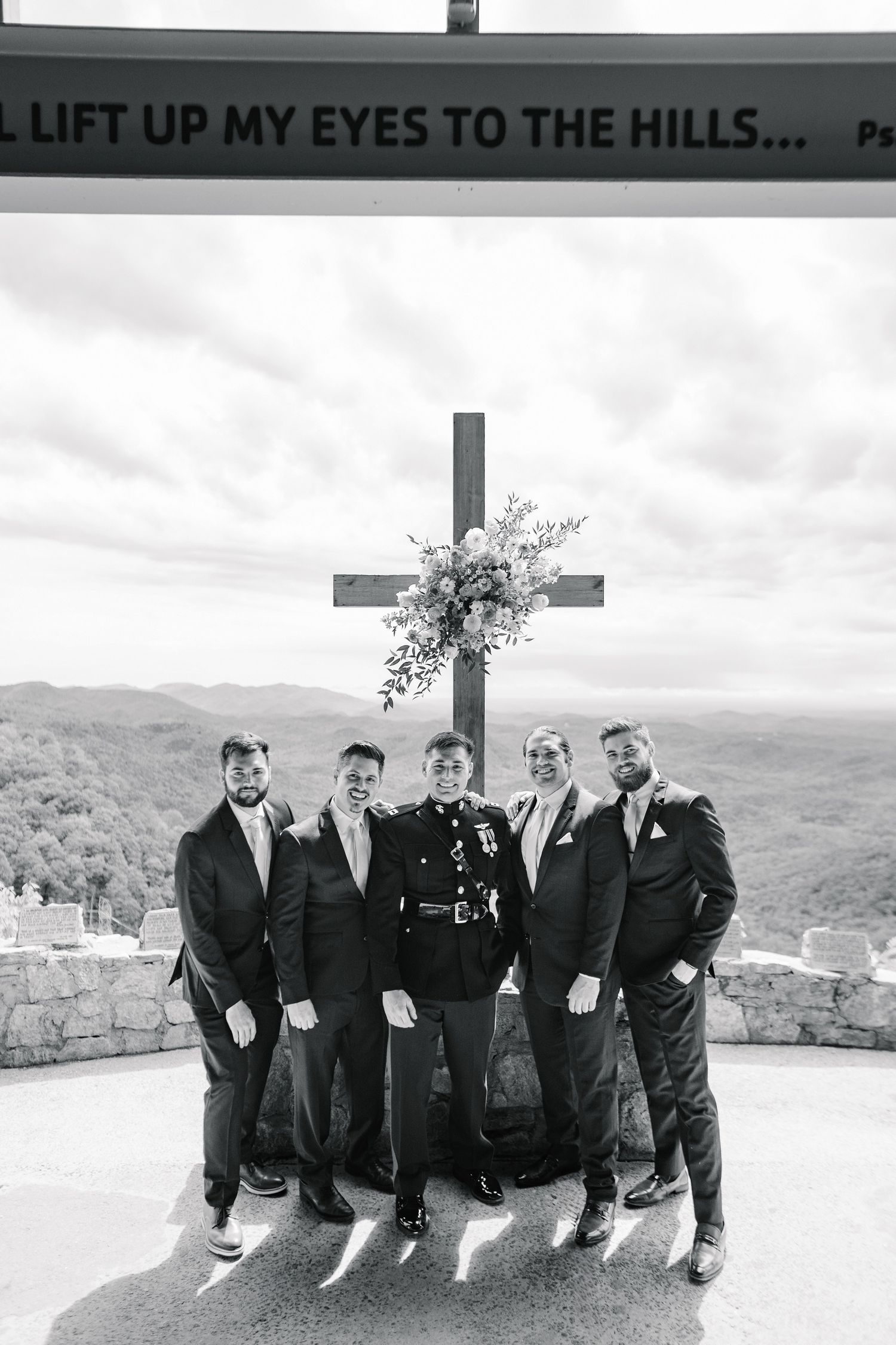 Black and white photograph of groomsmen standing beneath a cross and sign at a scenic overlook with mountain views.