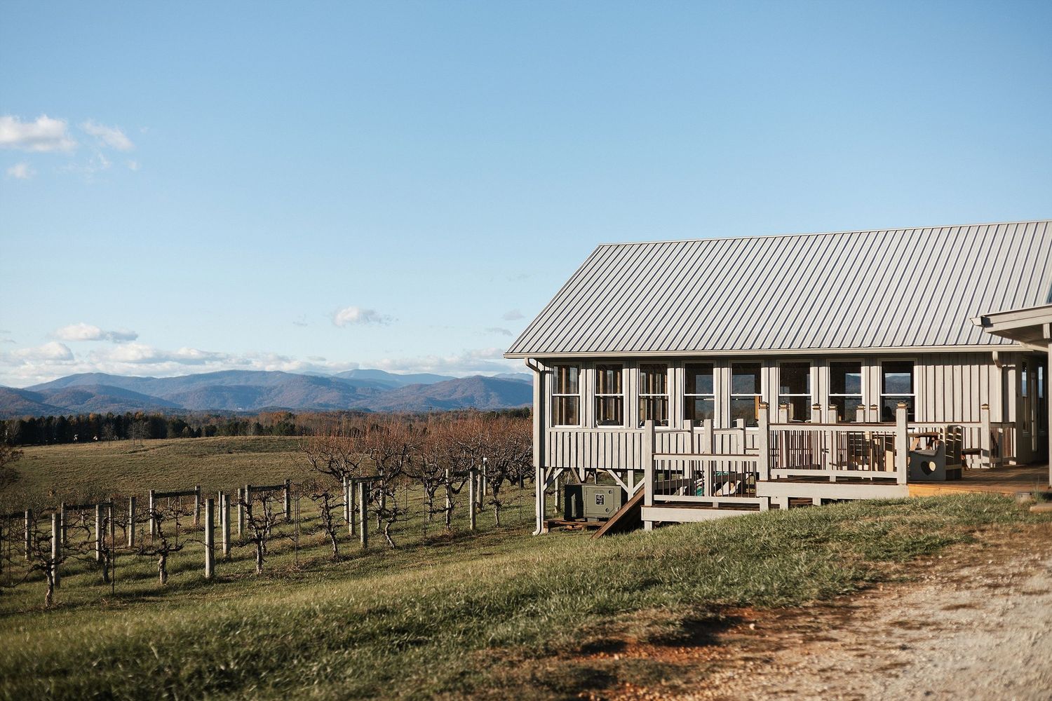 A rustic farmhouse with a wraparound porch overlooks rolling hills and vineyards under a blue sky.