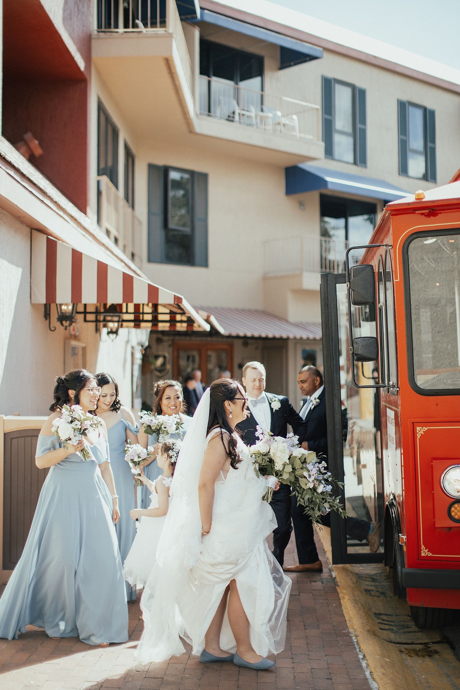 Wedding party poses outside building near red trolley in light blue bridesmaid dresses.