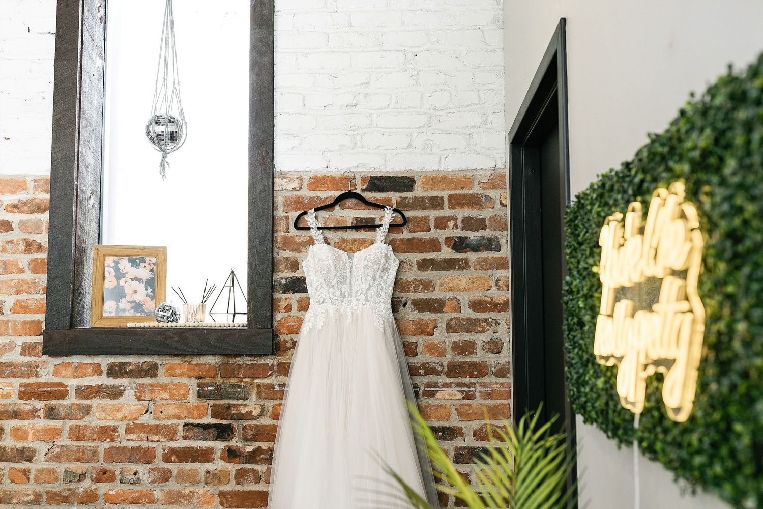An elegant white wedding dress hangs against exposed brick wall with a decorative mirror and greenery wreath.