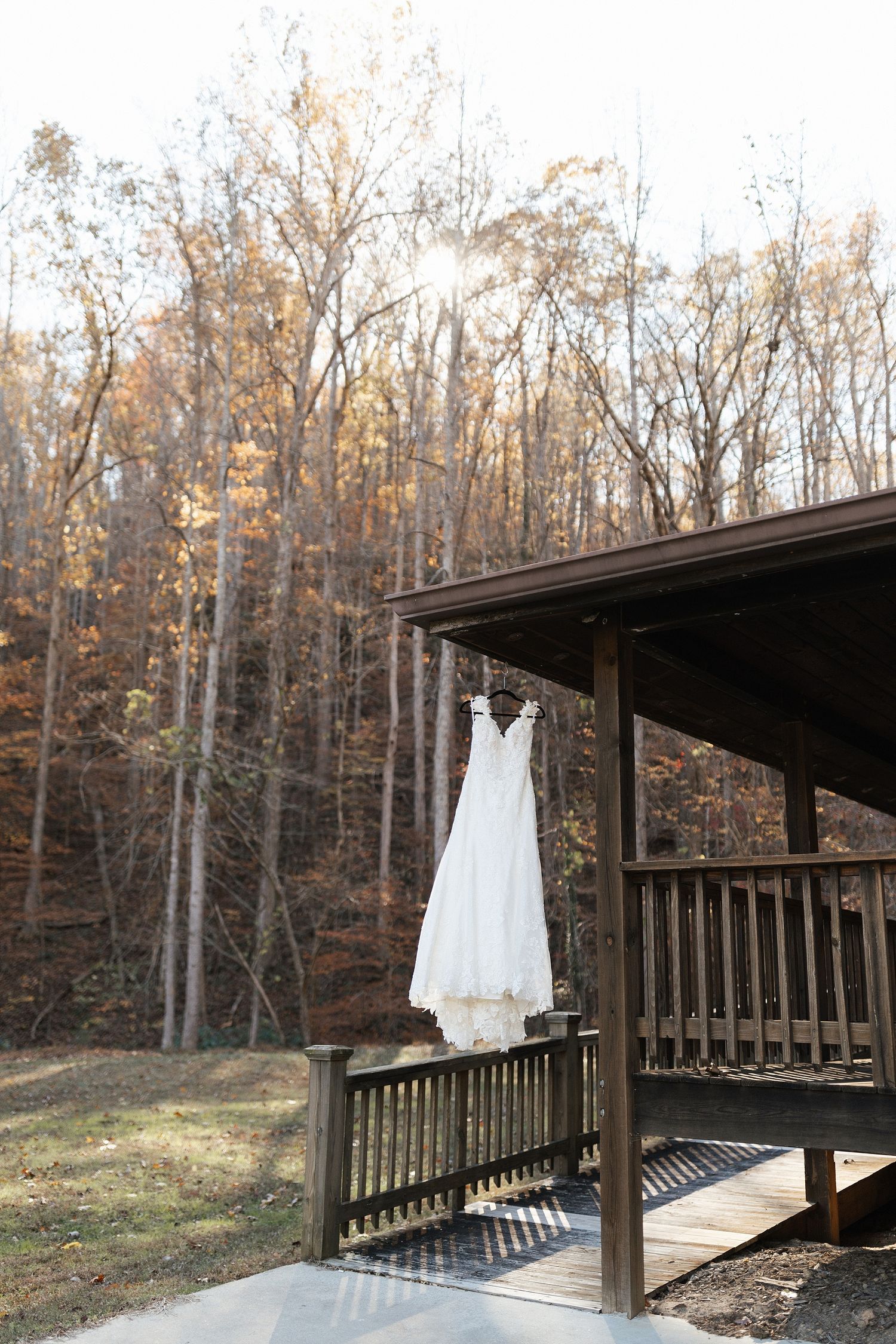 A white wedding dress hangs from a rustic wooden porch railing against an autumn forest backdrop.