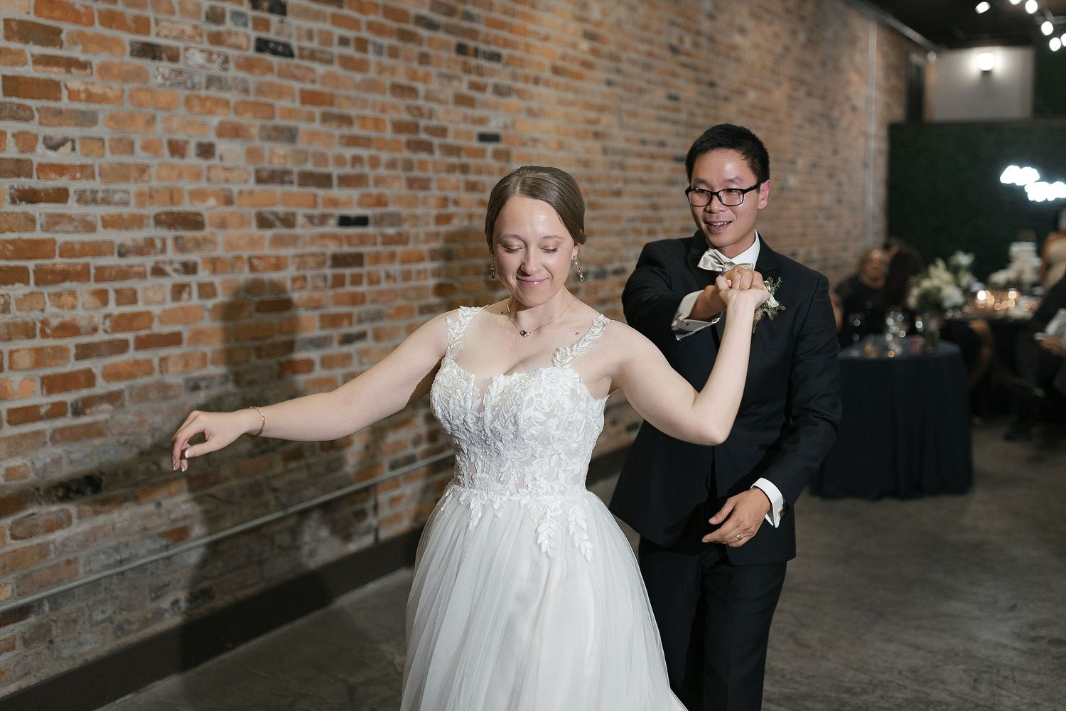 A joyful couple dances together at their wedding reception in front of an exposed brick wall.
