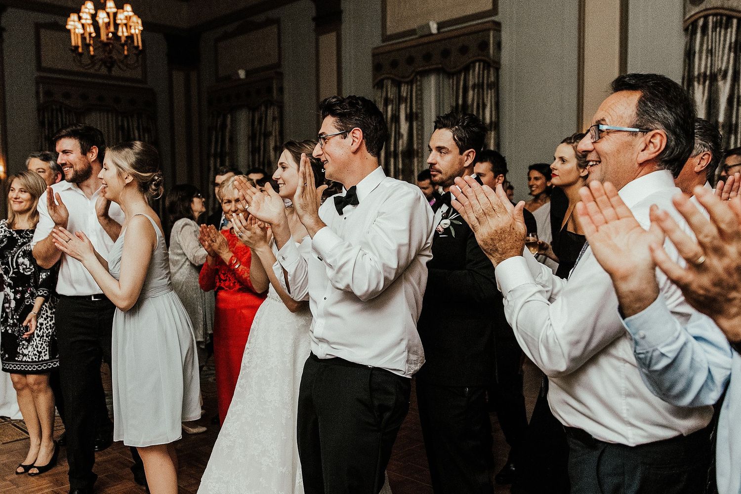 Wedding guests clapping and dancing in formal attire under a crystal chandelier.
