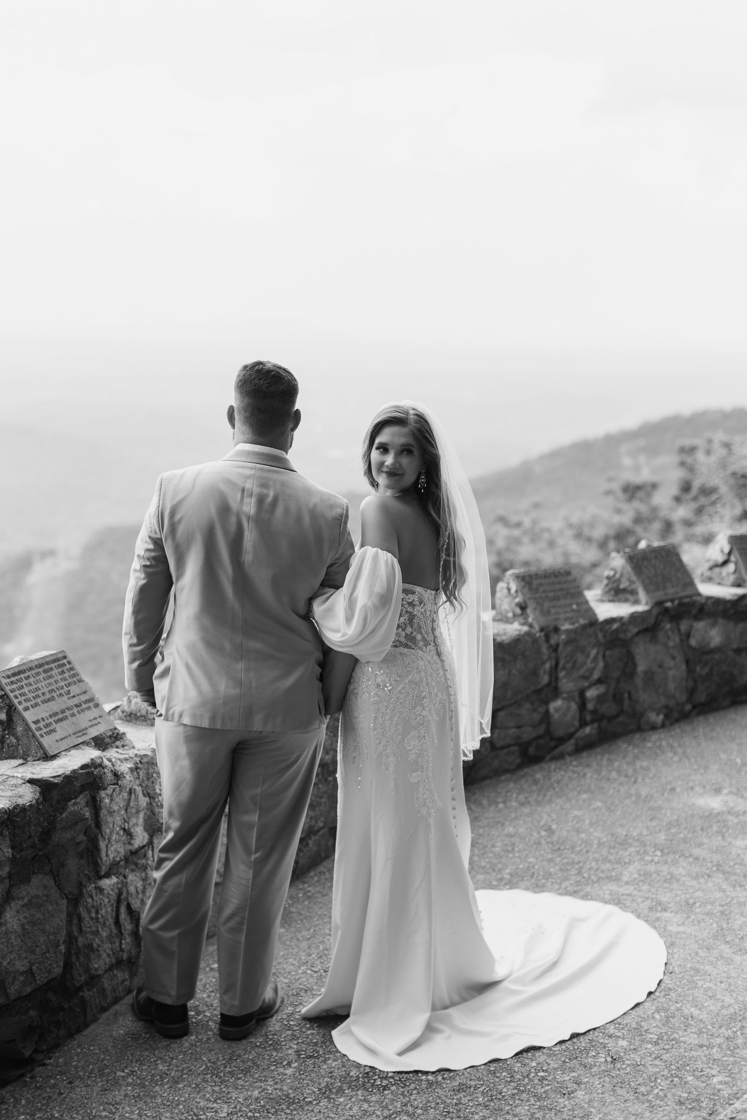 A black and white wedding photo of a couple standing at a scenic overlook with a stone wall and mountains in the background.