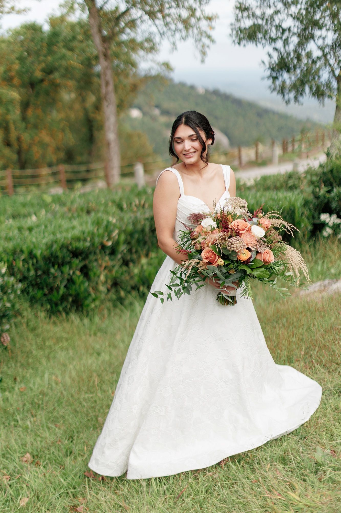 Bride in white lace gown holding lush peach and green bouquet, smiling outdoors with mountain backdrop.