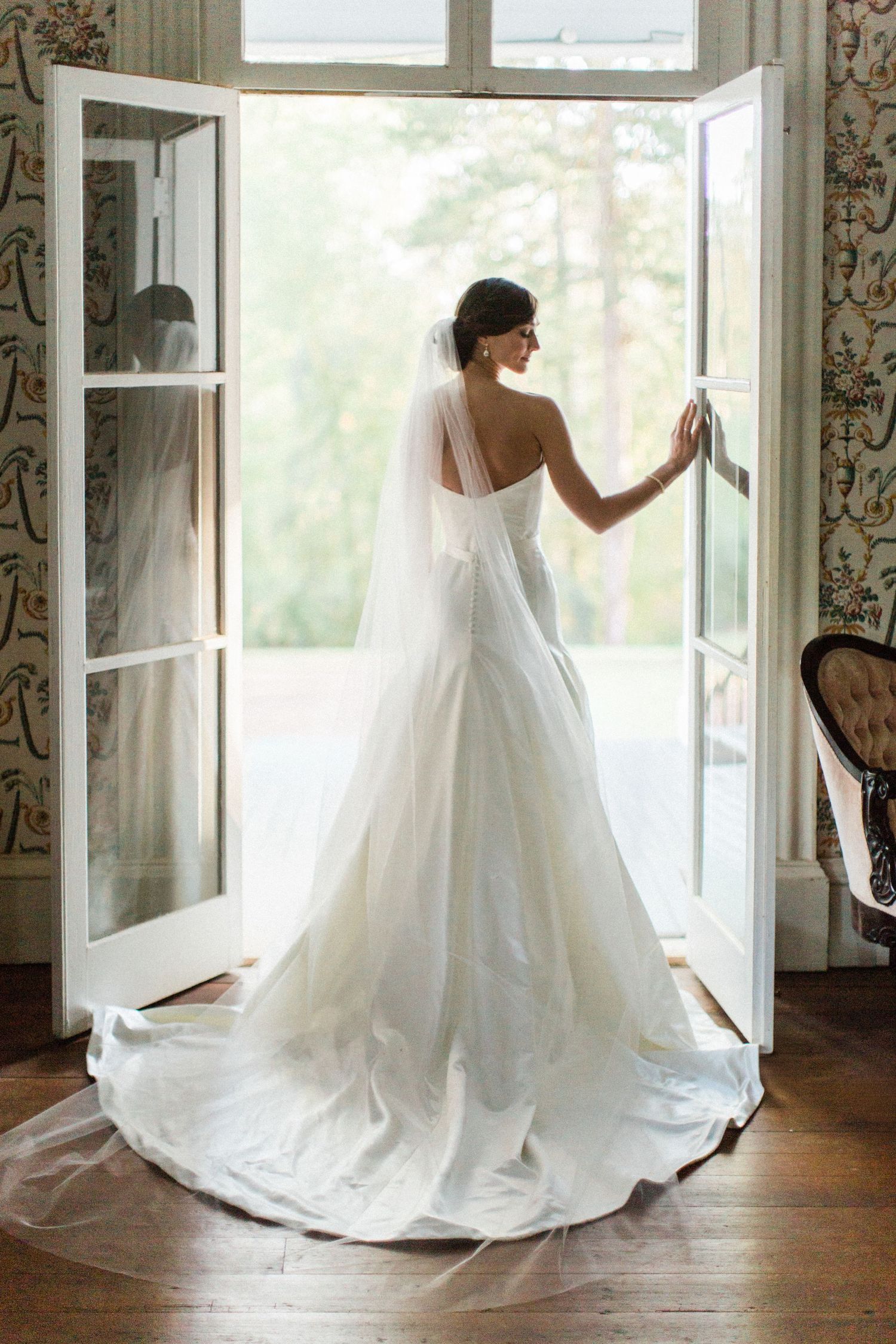 Bride in elegant white A-line wedding gown with long veil stands at open French doors, viewed from behind.