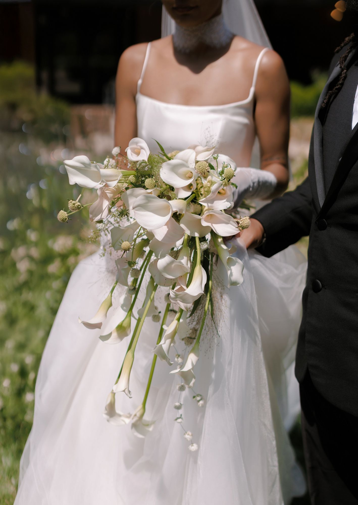 Bride in white gown holding cascading calla lily bouquet with greenery beside groom in black suit.