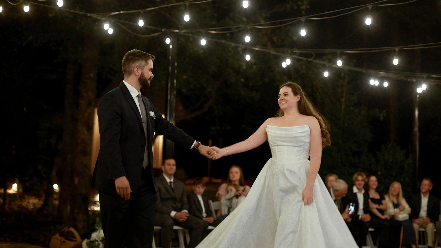 Bride and groom share first dance outdoors at night under string lights at elegant wedding reception.
