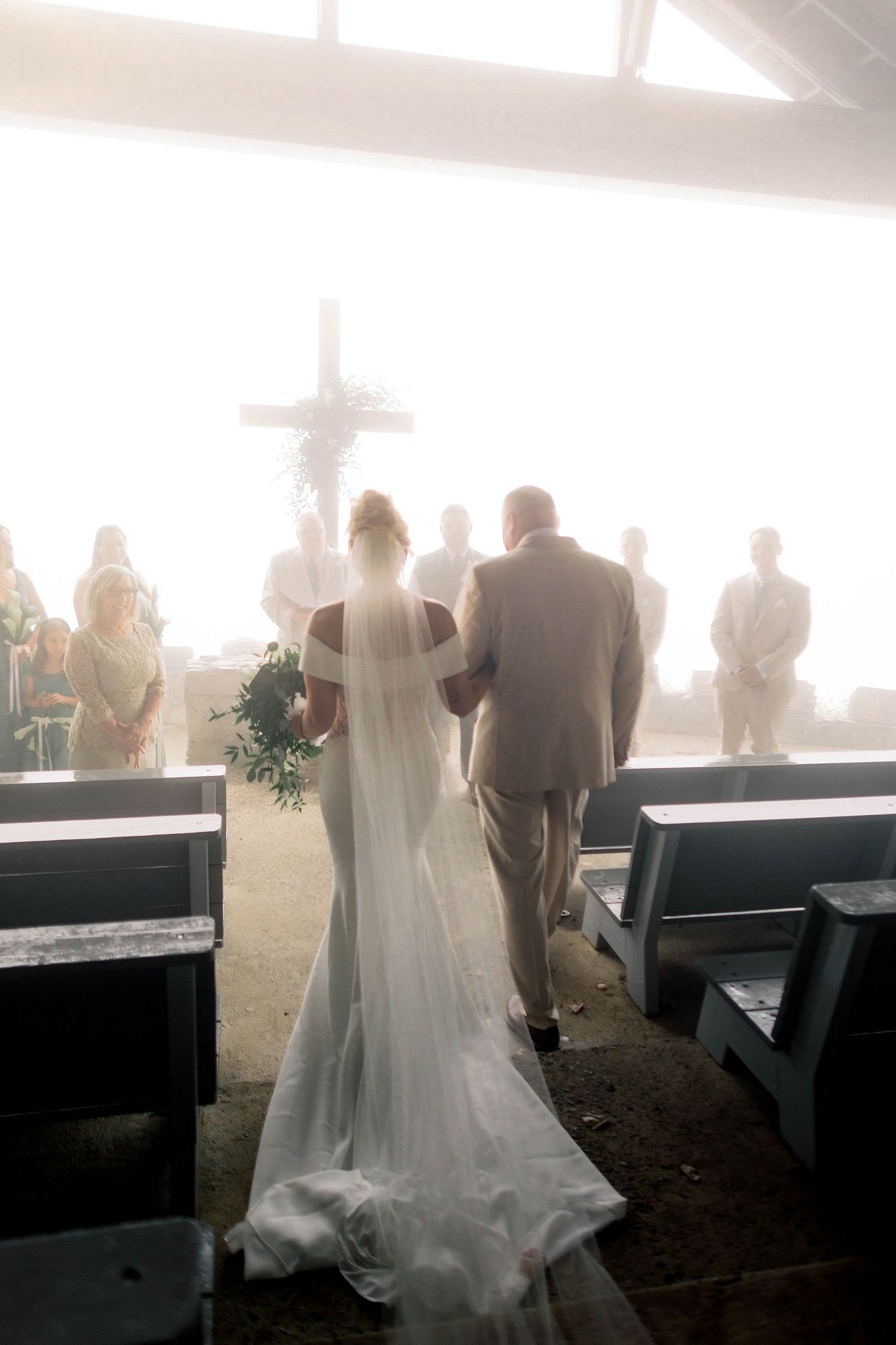 Bride walks down the aisle with escort toward a cross altar in a misty open-air mountain chapel.