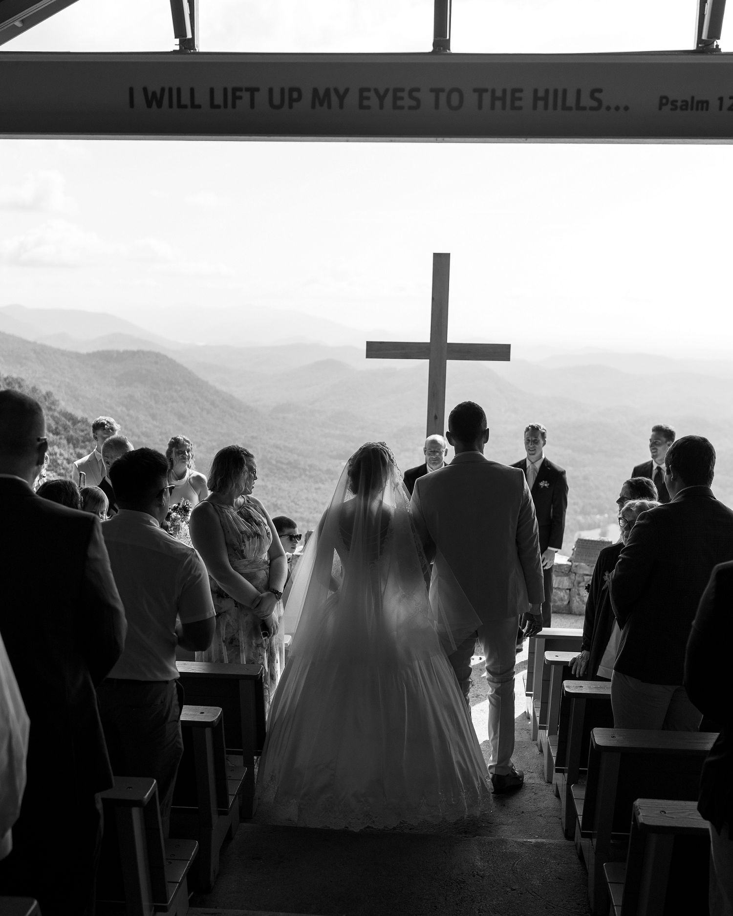 Black and white wedding ceremony photo inside a hilltop chapel with a cross and mountain views.