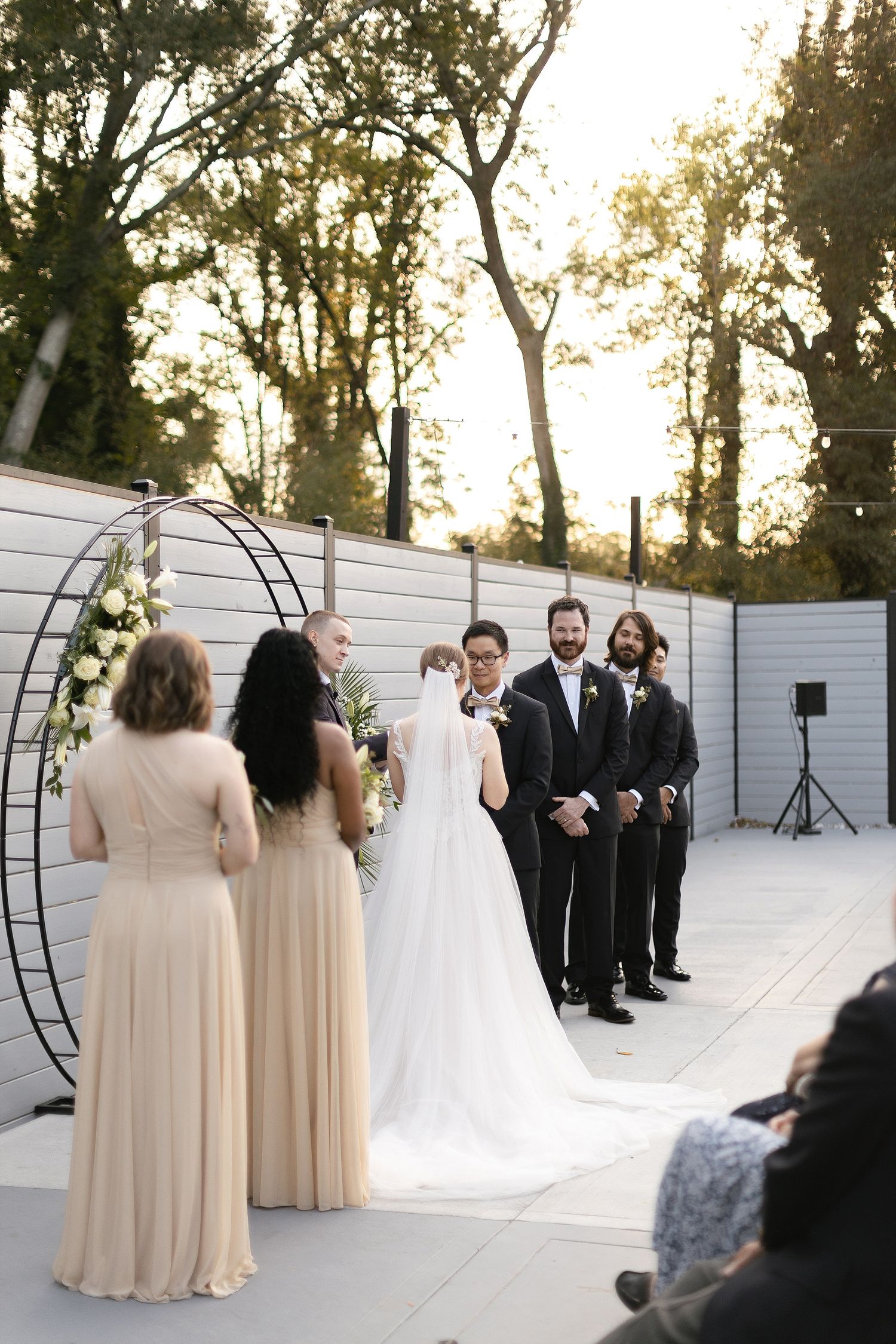 Wedding party stands before a modern circular arch with white floral arrangements against a gray wall backdrop at sunset.
