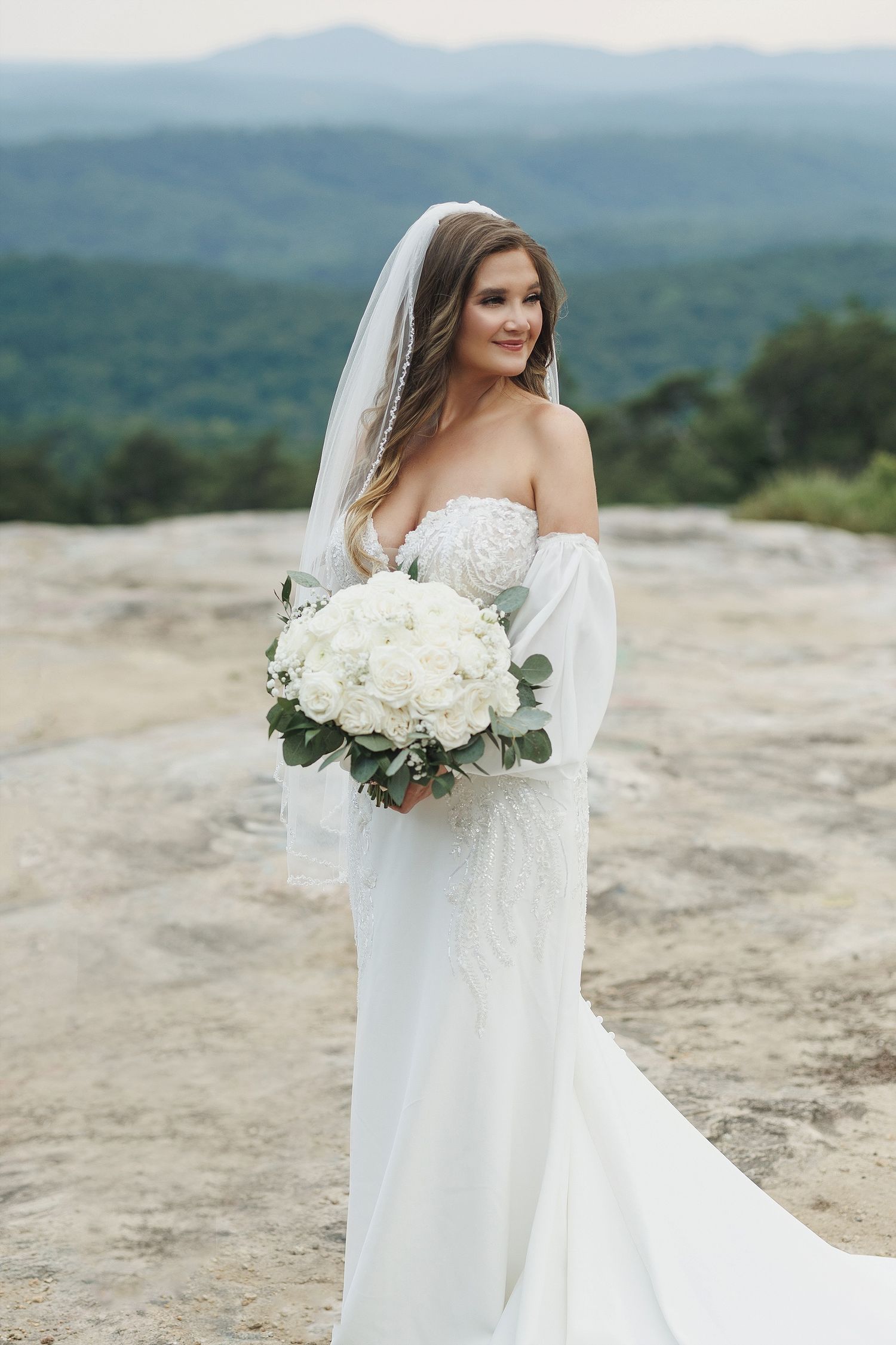 Bride in off-shoulder gown holds white bouquet against mountain landscape backdrop.