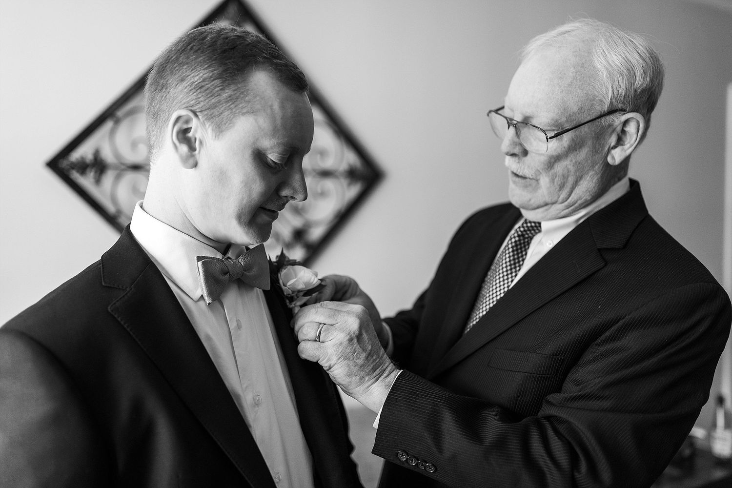 A tender moment captured in black and white as an older gentleman helps adjust a bow tie on a younger man in formal attire.