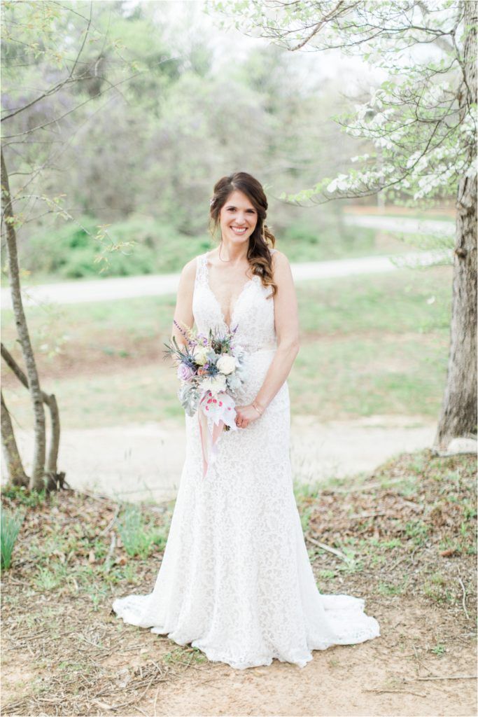 Bride in lace gown holds pastel bouquet while standing among spring trees.