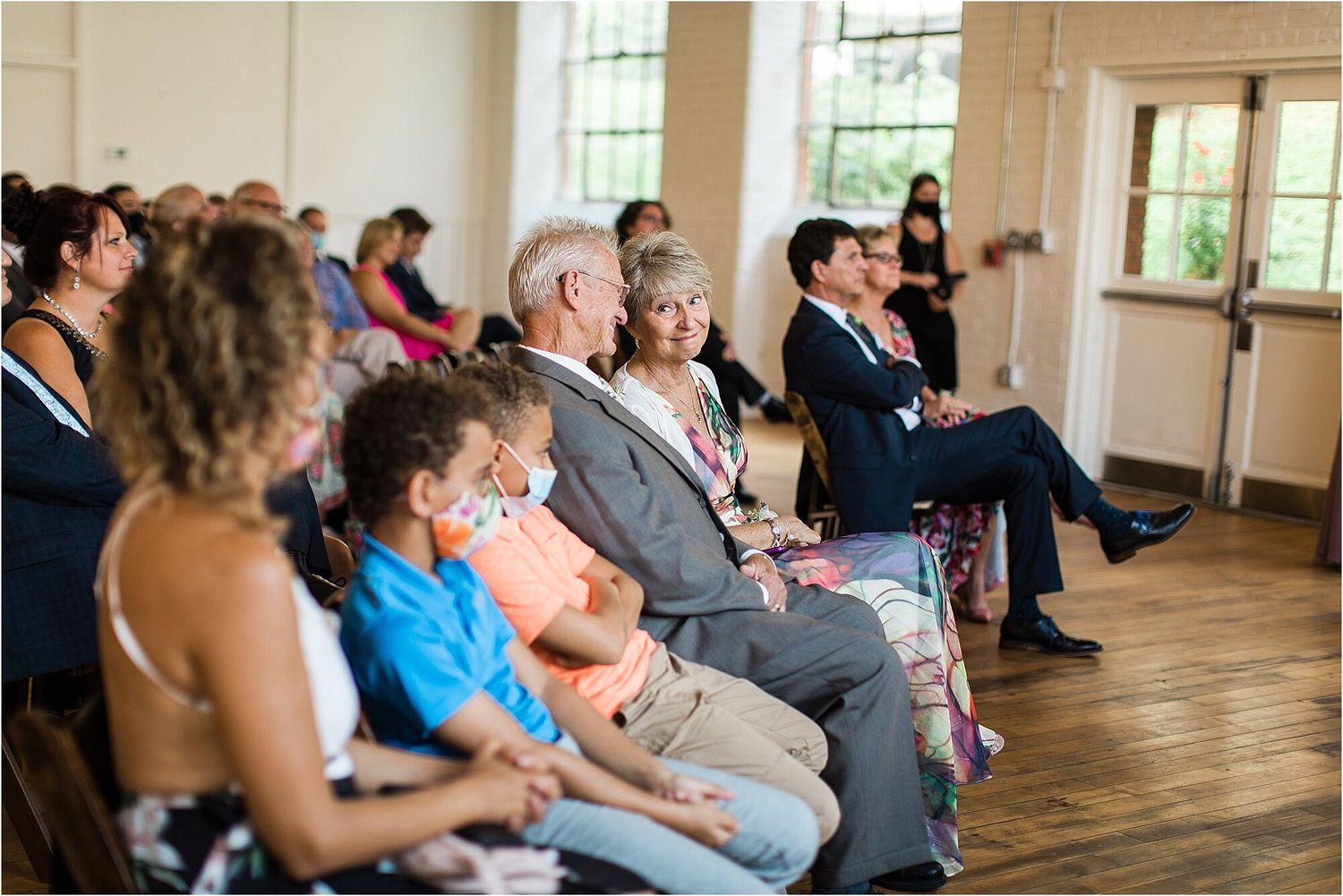 Wedding guests seated in rows watch the ceremony in a bright indoor venue with hardwood floors.