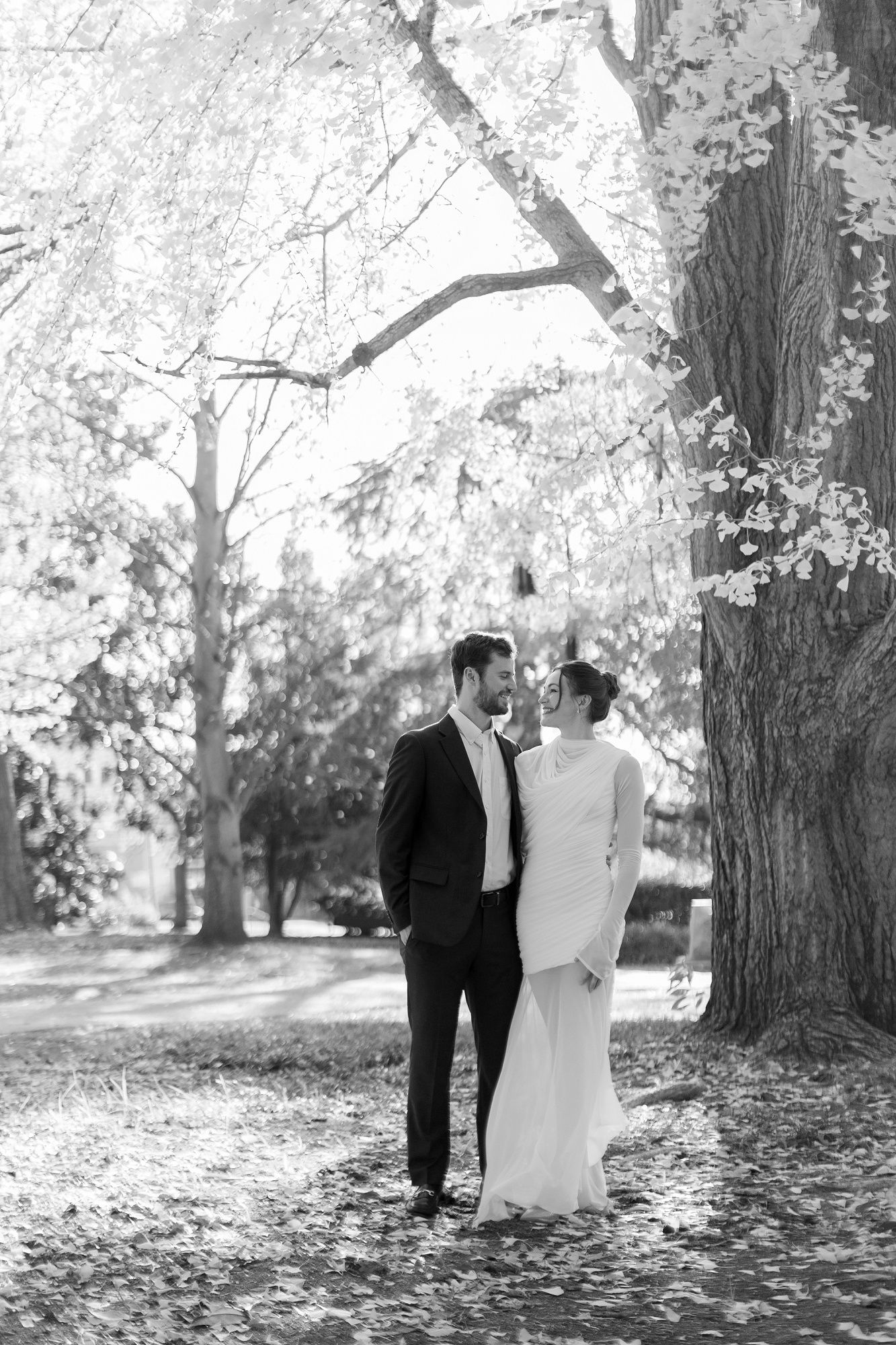 A romantic couple walks together through a park filled with autumn leaves and trees in black and white photography.