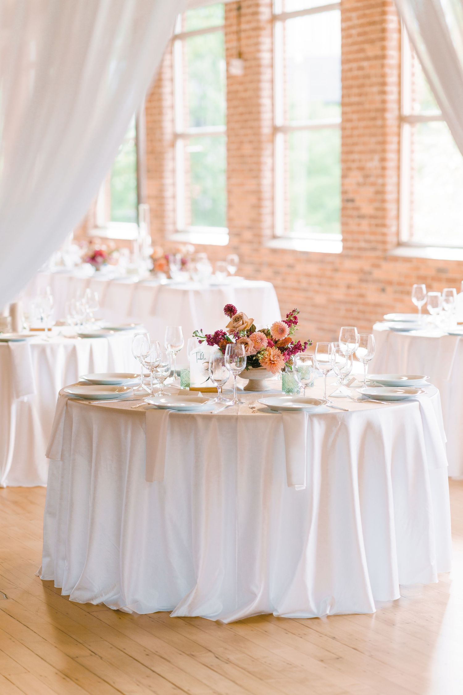 Elegant wedding reception tables with white draping, colorful floral centerpieces, and gold accents in a brick venue.