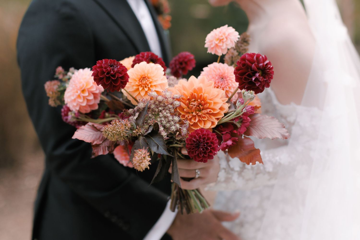 Bride and groom holding a lush fall wedding bouquet featuring orange, pink, and burgundy dahlias.