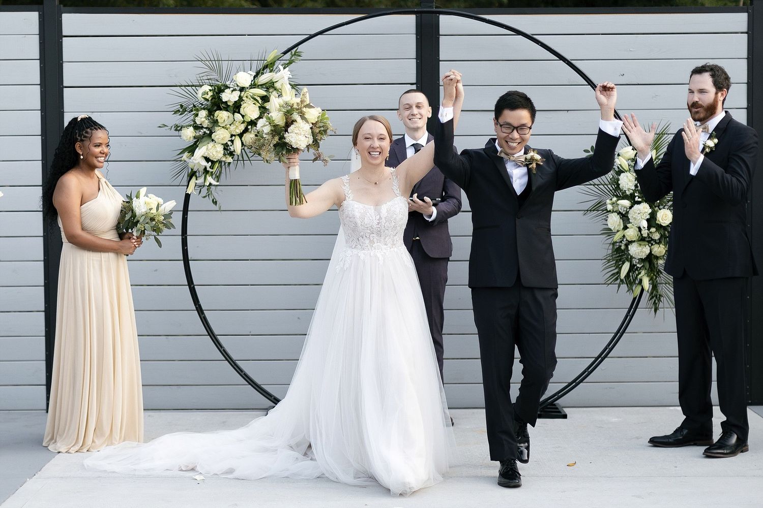 Wedding party celebrates by circular arch decorated with white floral arrangements against gray wall.
