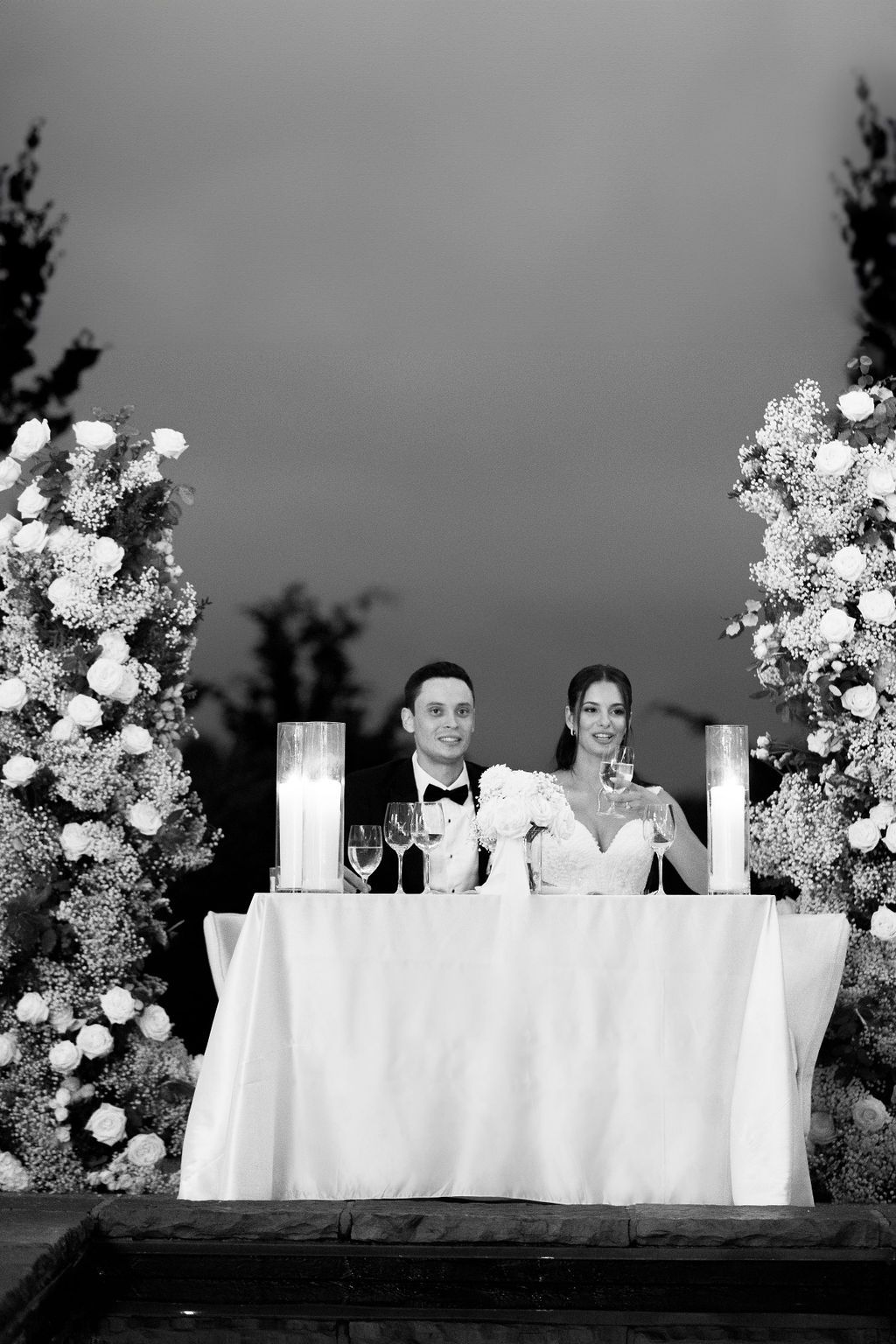 Black and white wedding photo of couple at sweetheart table with floral arch arrangements.