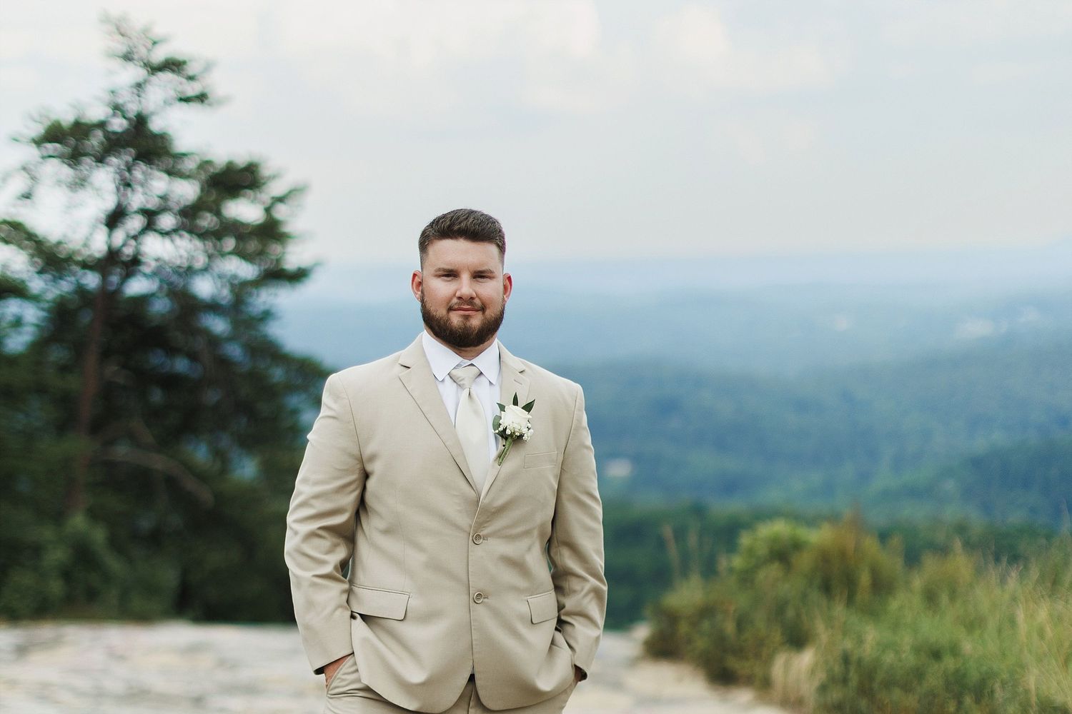 A groom in beige suit stands on rocky mountain overlook with scenic valley view.