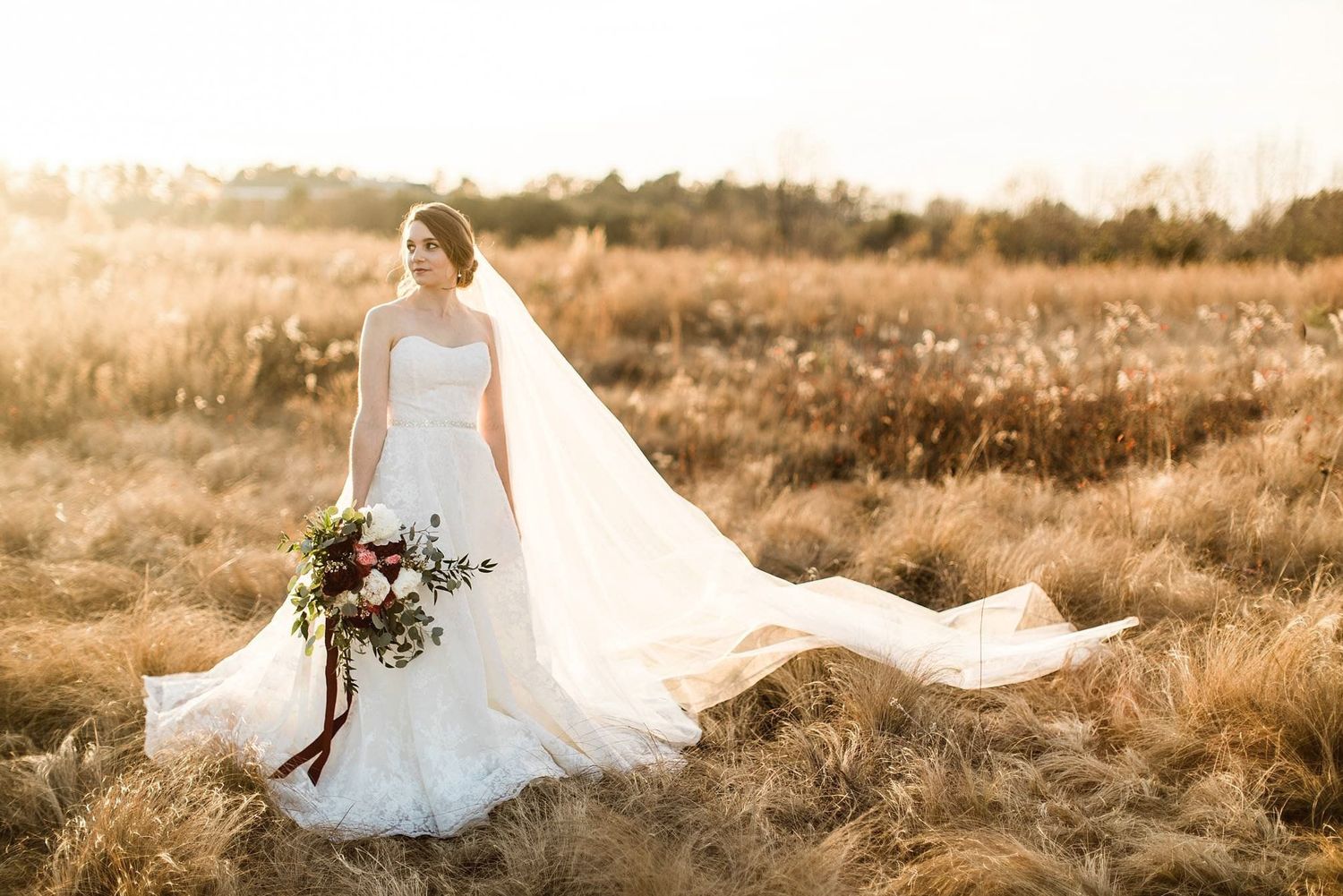 A bride in a flowing white gown and veil holds a burgundy bouquet while standing in a golden sunlit field at sunset.