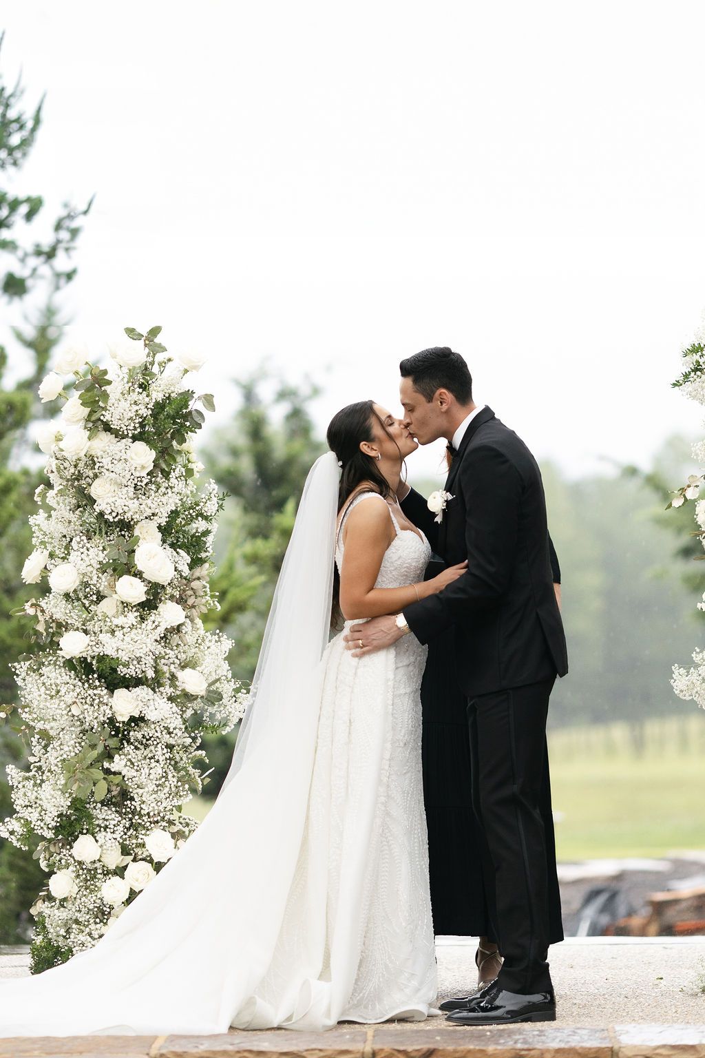 Romantic wedding moment under white floral arch in outdoor setting.
