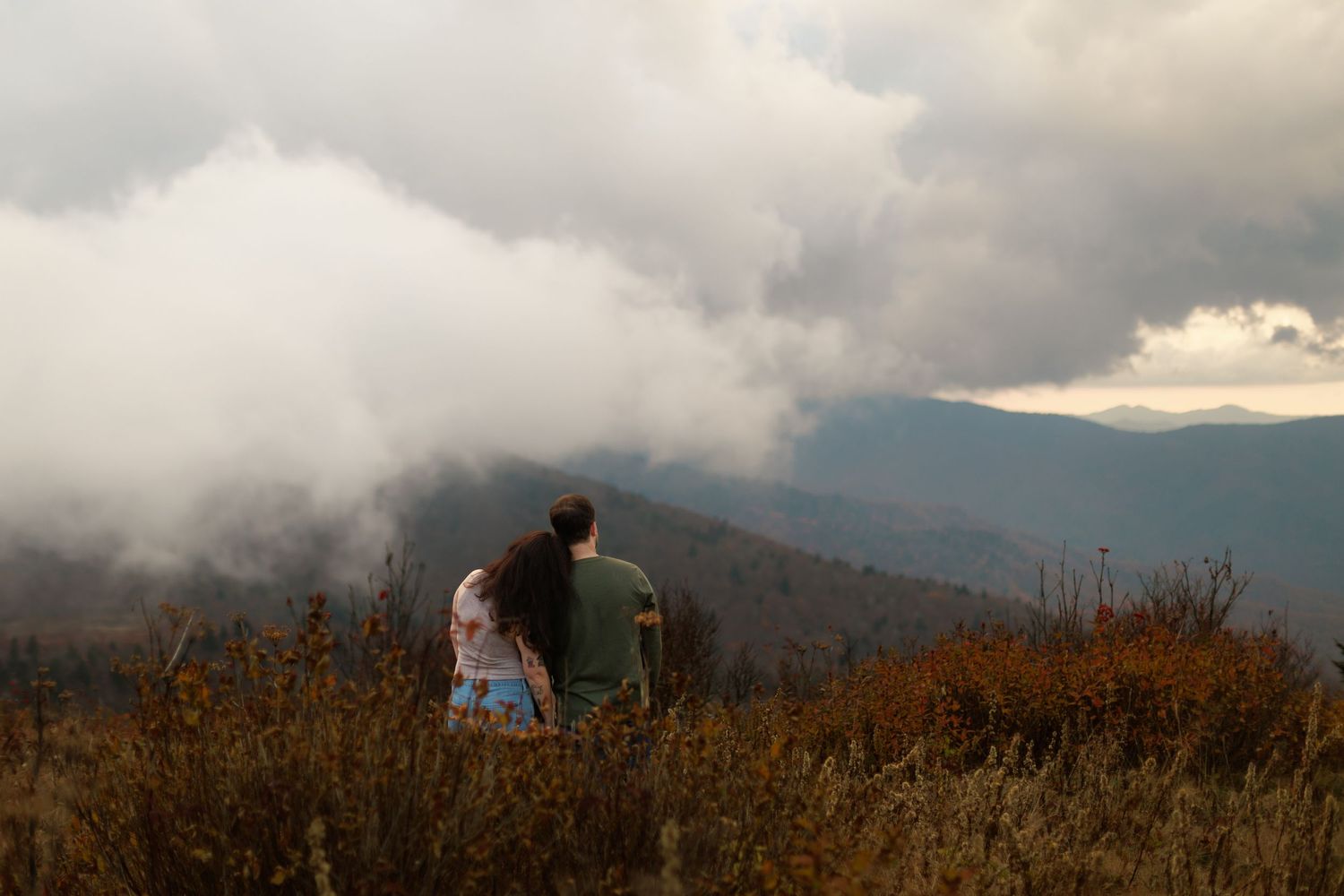 A panoramic mountain landscape with misty clouds rolling over rugged peaks and brown autumn grass in the foreground.