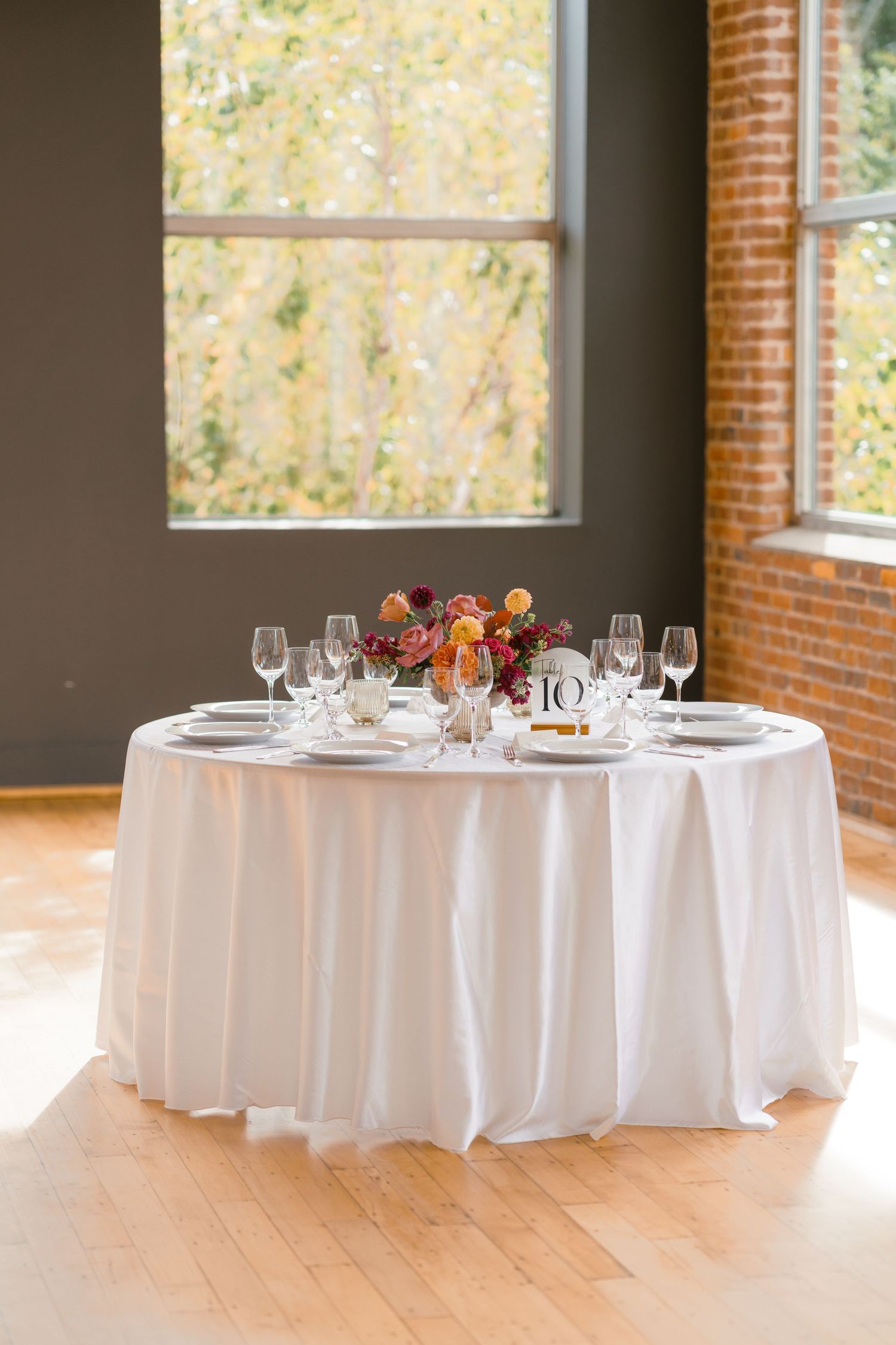Elegant round wedding reception table with white linen, crystal glasses, and a colorful floral centerpiece by a window.