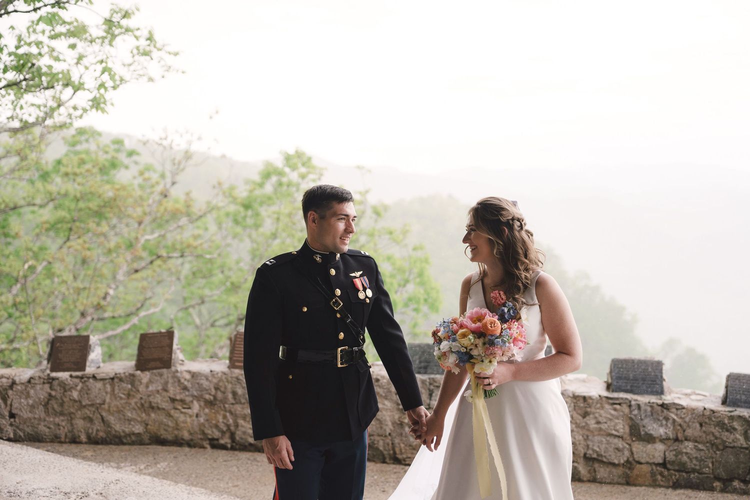 Romantic couple in wedding attire share a playful moment walking together outdoors on a sunlit path.