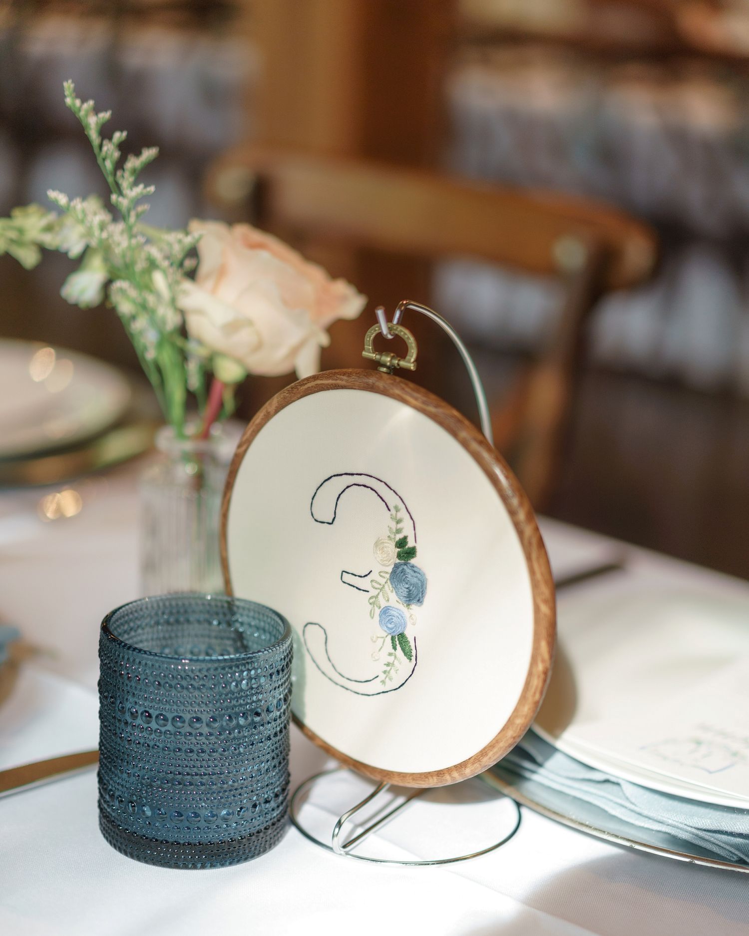 Elegant wedding table number 3 in embroidery hoop with floral design, blue textured candle holder, and pink roses centerpiece.