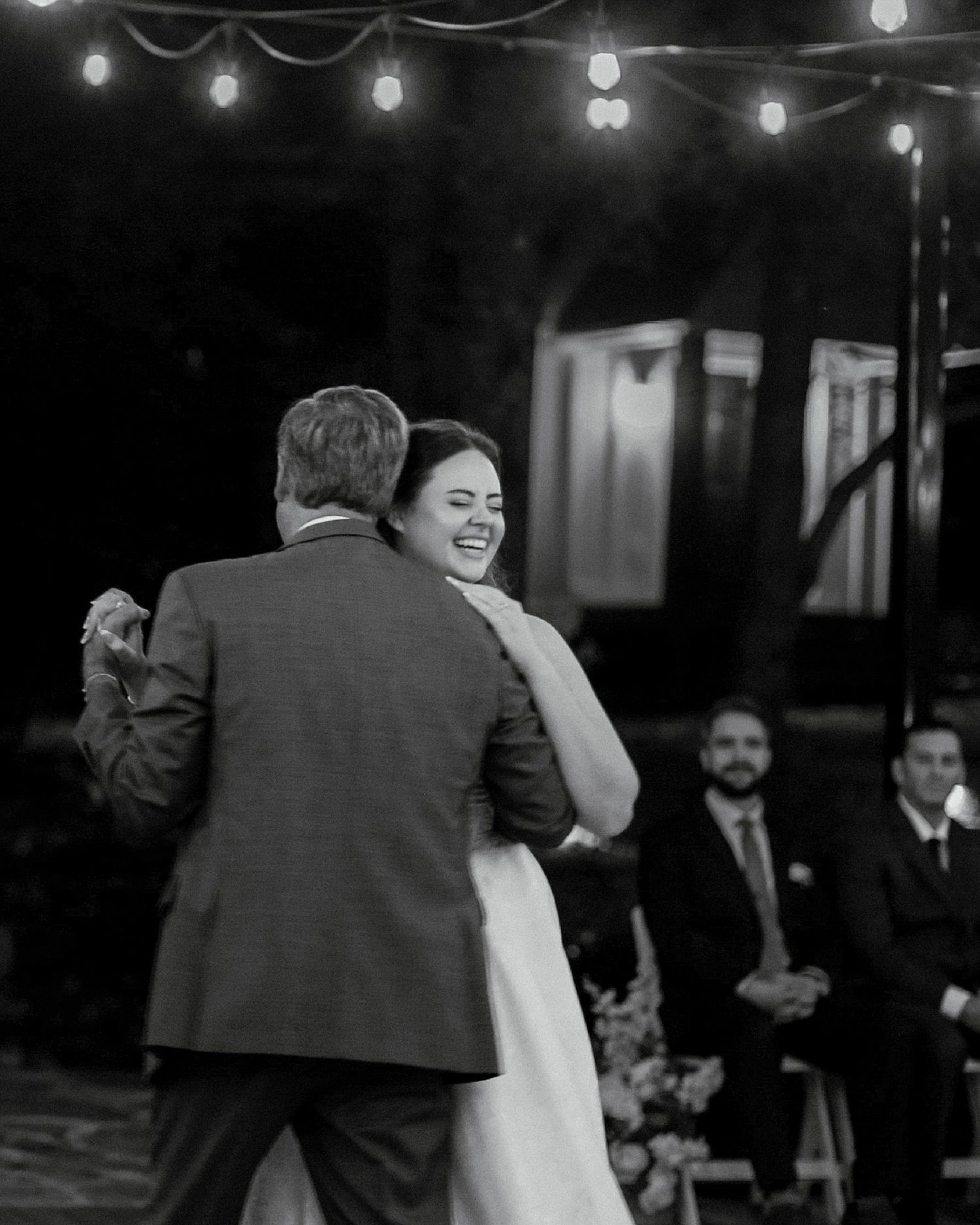 Black and white photo of a bride laughing while sharing a dance outdoors under string lights at her wedding reception.