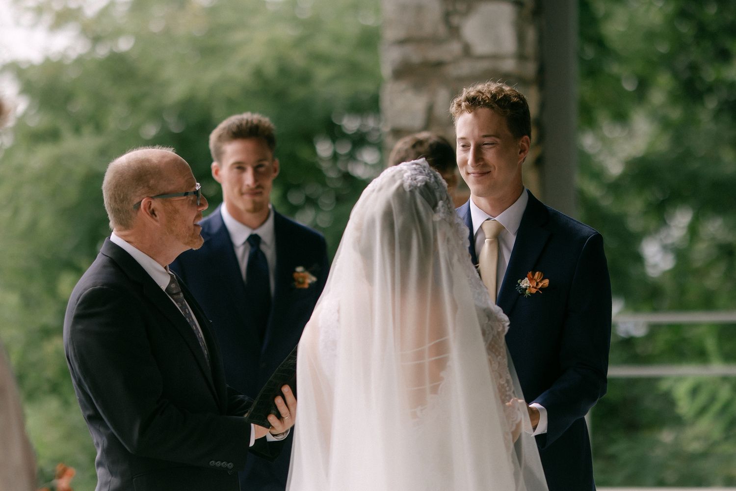 Groom smiling at veiled bride during outdoor wedding ceremony with officiant and groomsman in background.