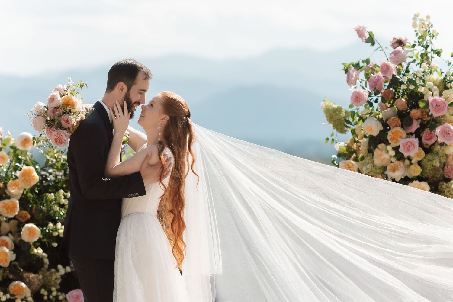 Bride and groom embrace at outdoor wedding ceremony with floral arrangements and mountain views backdrop.