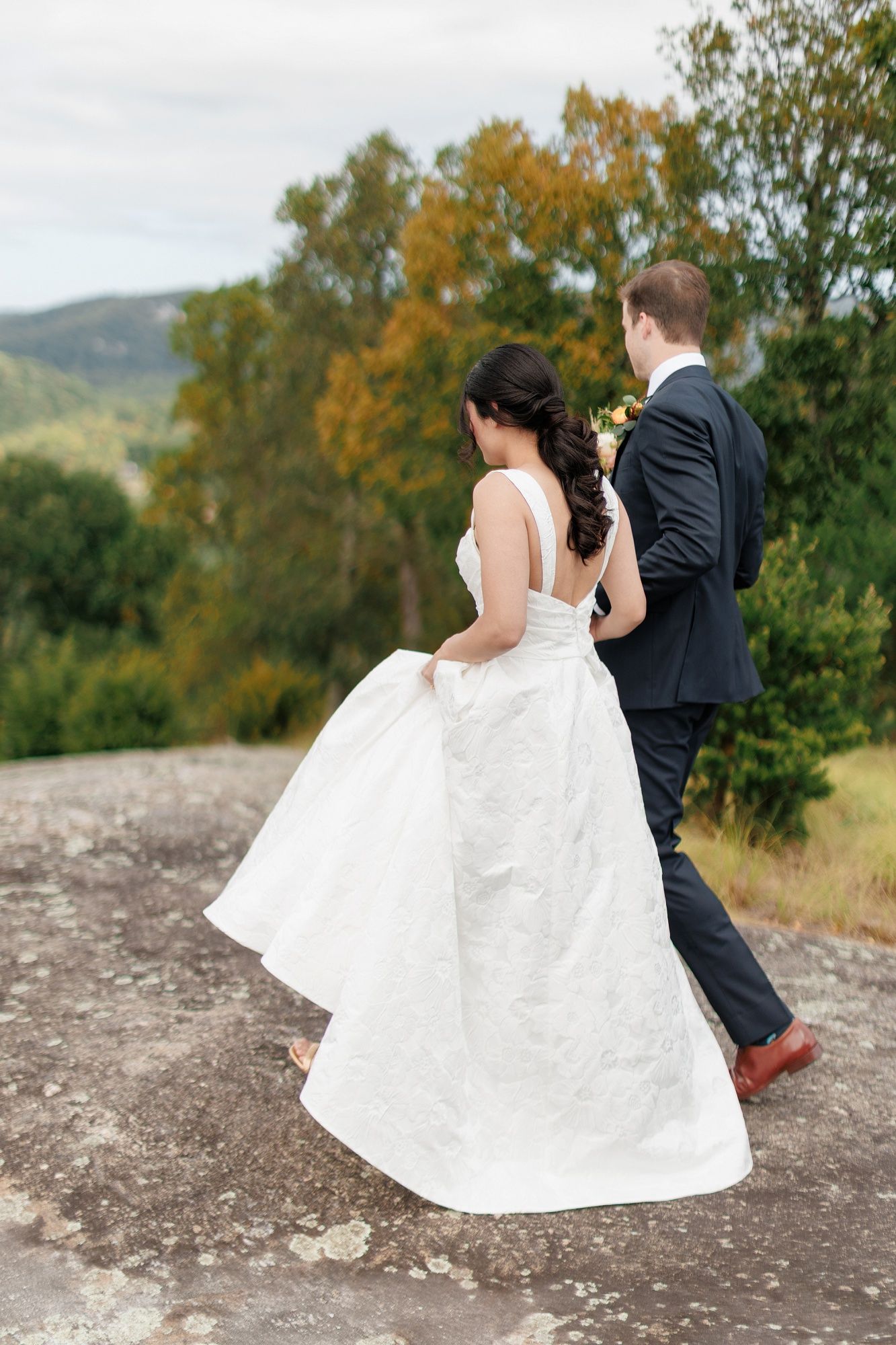Bride in white gown and groom in dark suit walk hand in hand on rocky overlook with lush green mountains behind.