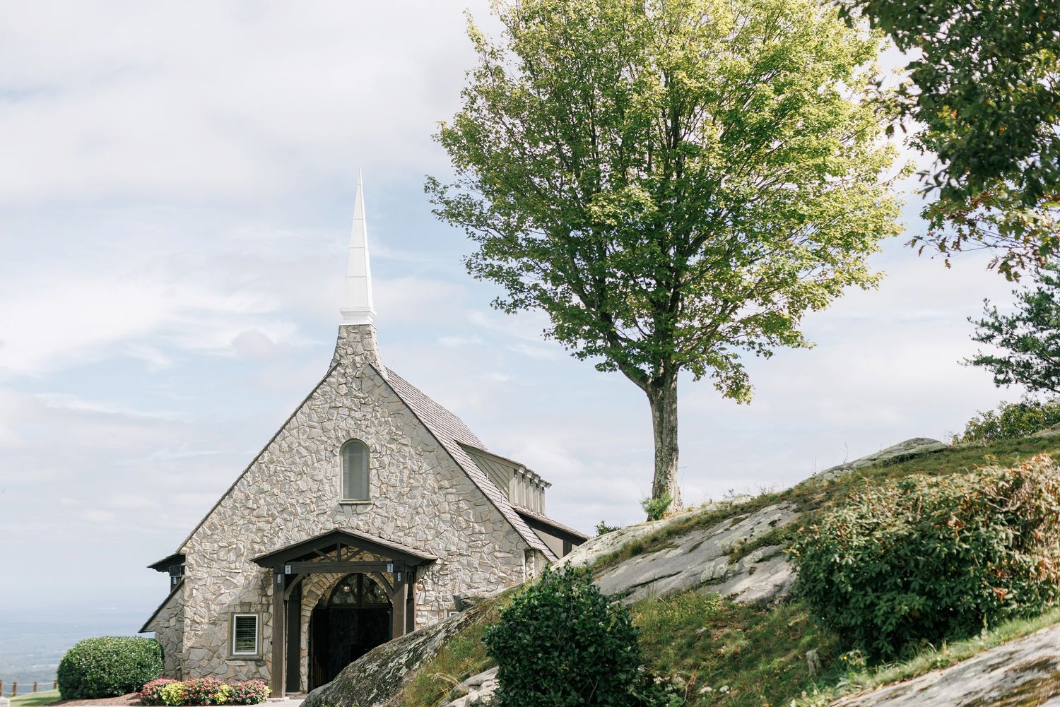 A stone chapel with a white steeple sits on a rocky hillside beside a tall tree under a cloudy sky.
