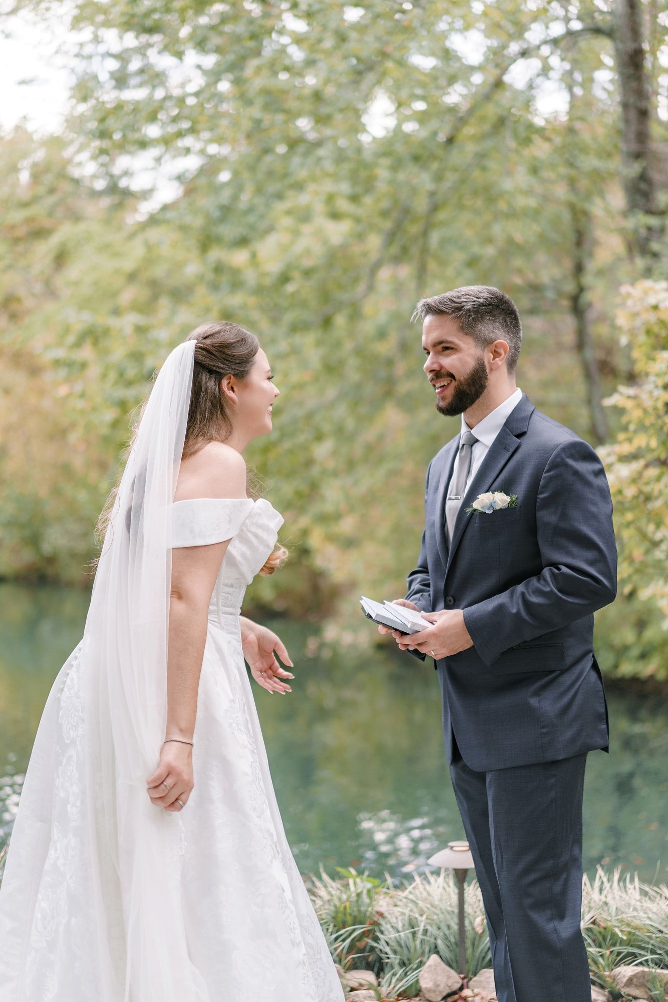 Bride in off-shoulder white gown and groom in navy suit exchange vows by a serene woodland pond.