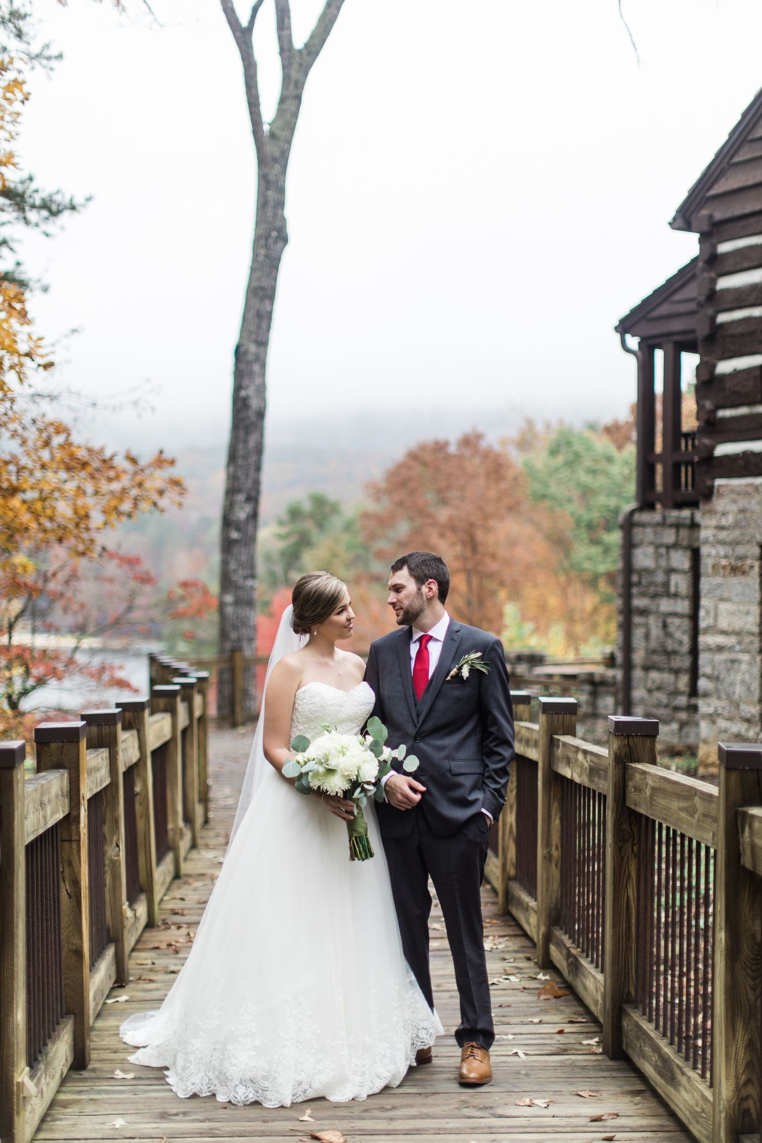 A couple stands together on a wooden walkway surrounded by autumn foliage on their wedding day.