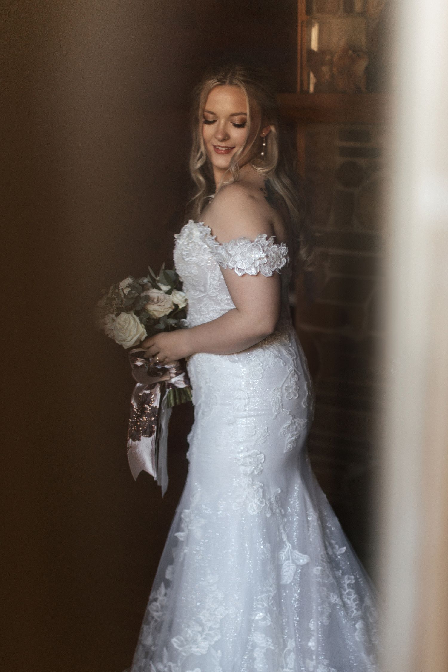 A bride in a fitted off-shoulder lace wedding gown holds a white bouquet while smiling in soft lighting.