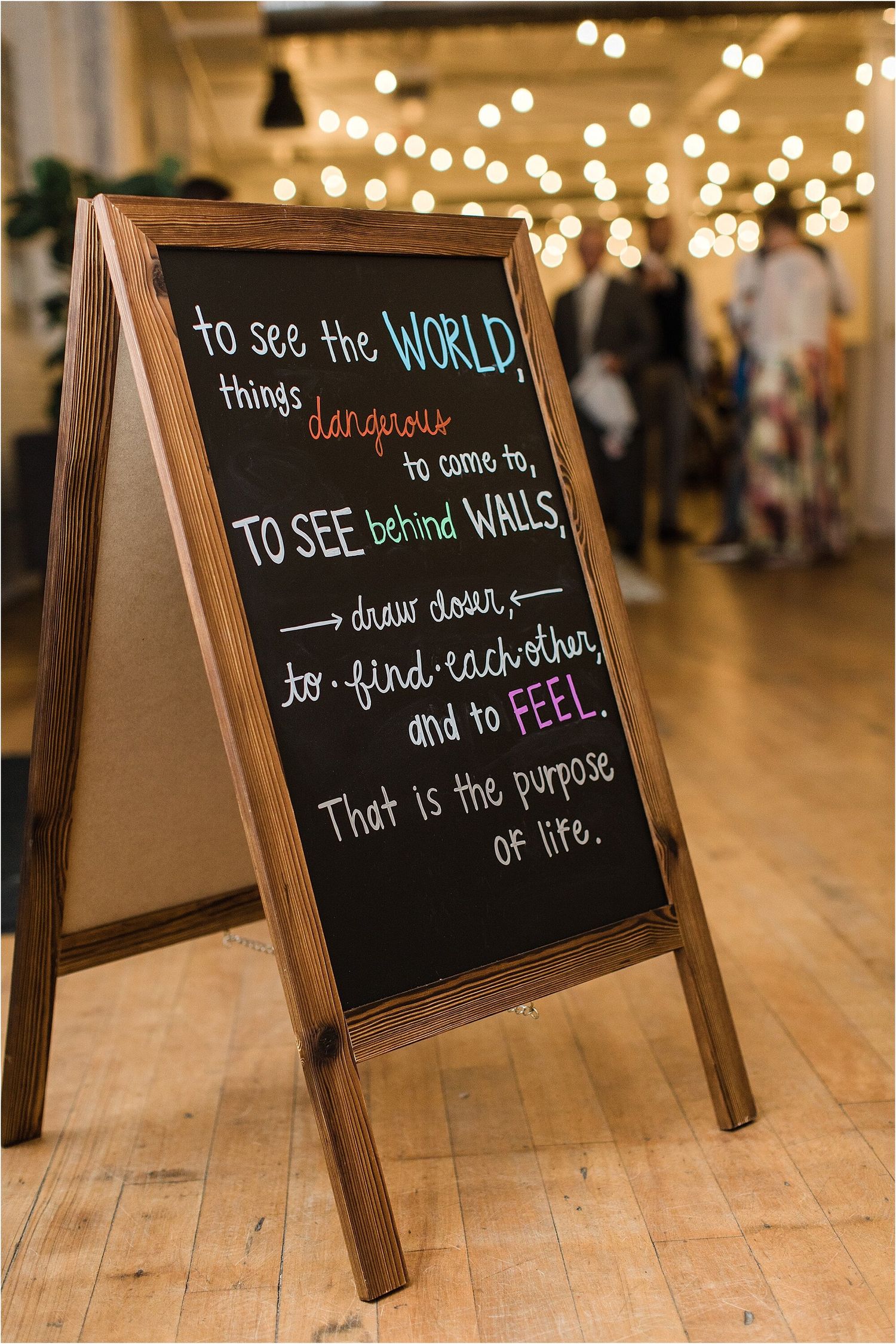 Rustic wooden chalkboard sign with inspirational quote about seeing the world, illuminated by bokeh lights in background.