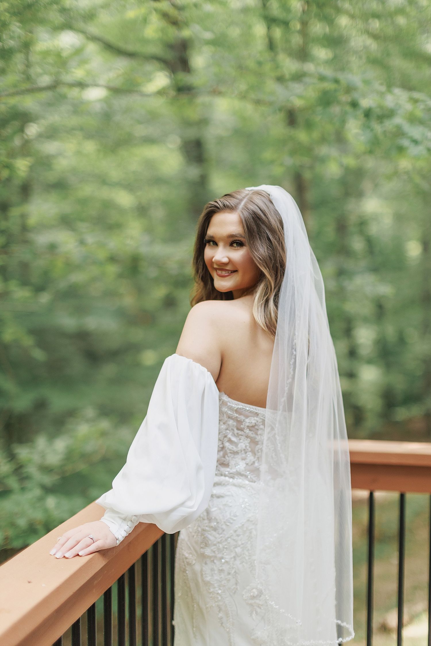 Bride in off-shoulder wedding dress poses on wooden deck with forest backdrop.