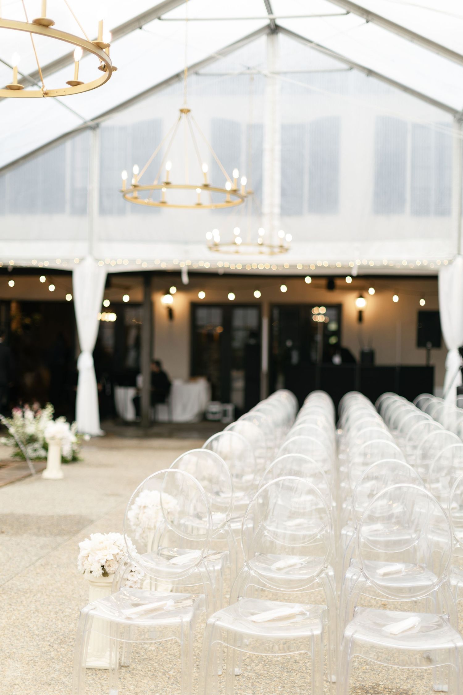 White outdoor wedding tent interior with hanging chandeliers and clear ghost chairs set up for an elegant ceremony.
