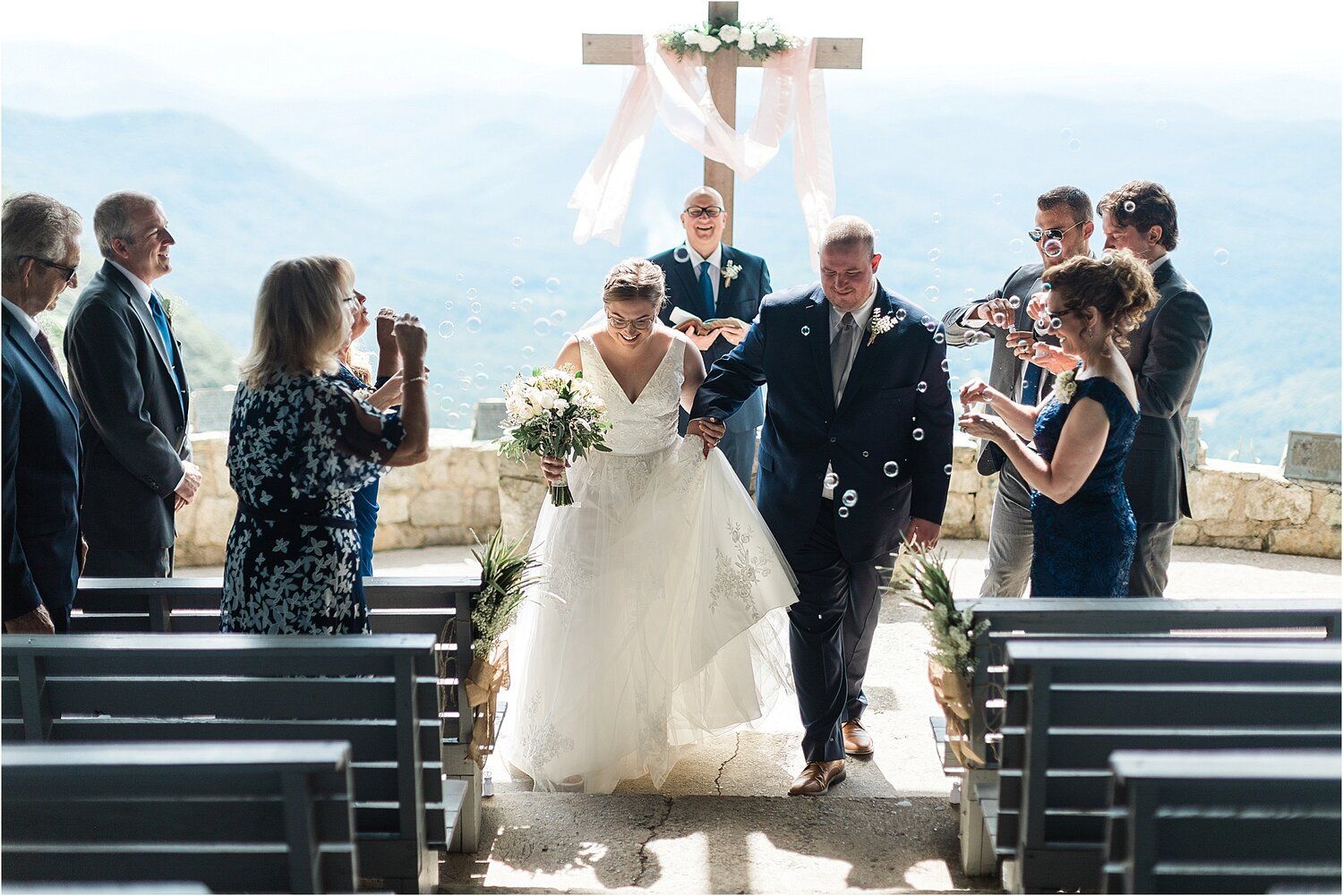 A wedding ceremony takes place outdoors with a mountain backdrop and crucifix as guests look on.