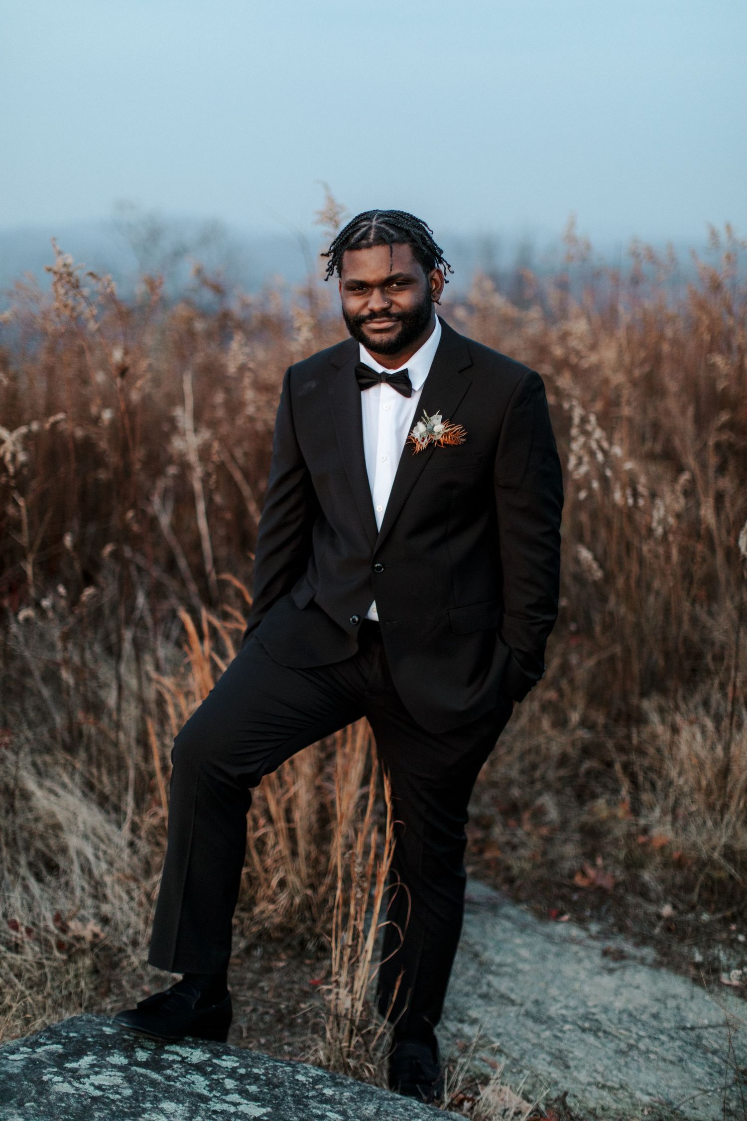 A groom in classic black tuxedo poses confidently against natural landscape.