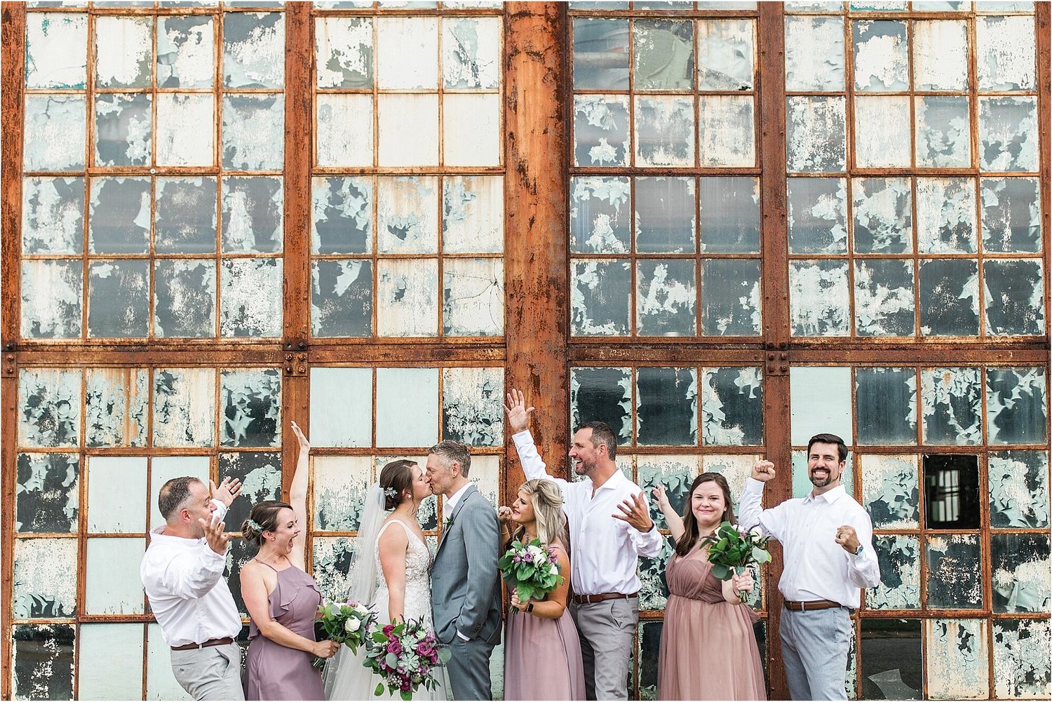 Bridal party celebrates with raised arms against weathered factory windows.