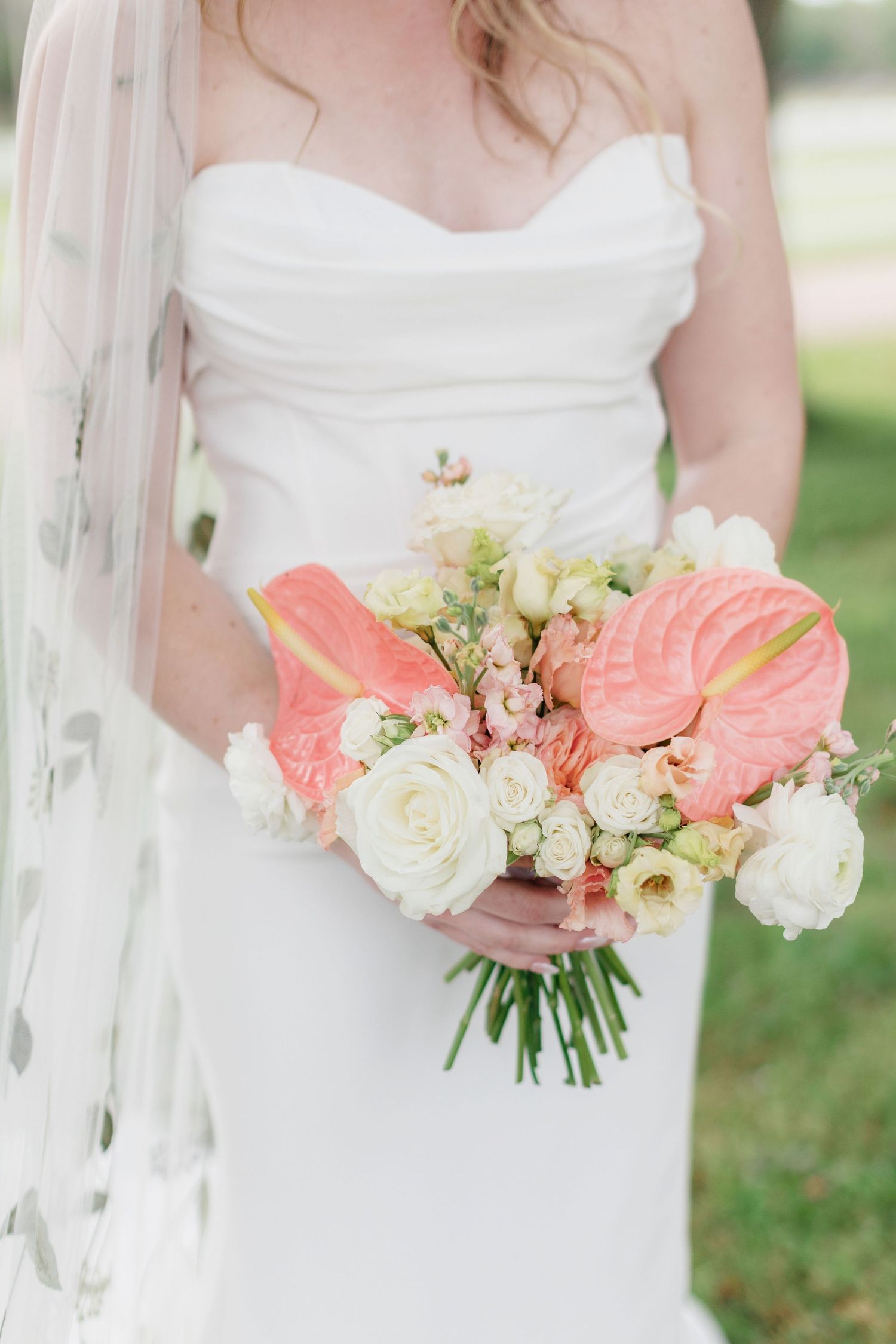 Elegant bridal bouquet of coral anthurium lilies, white roses and delicate greenery held against a classic white wedding gown.